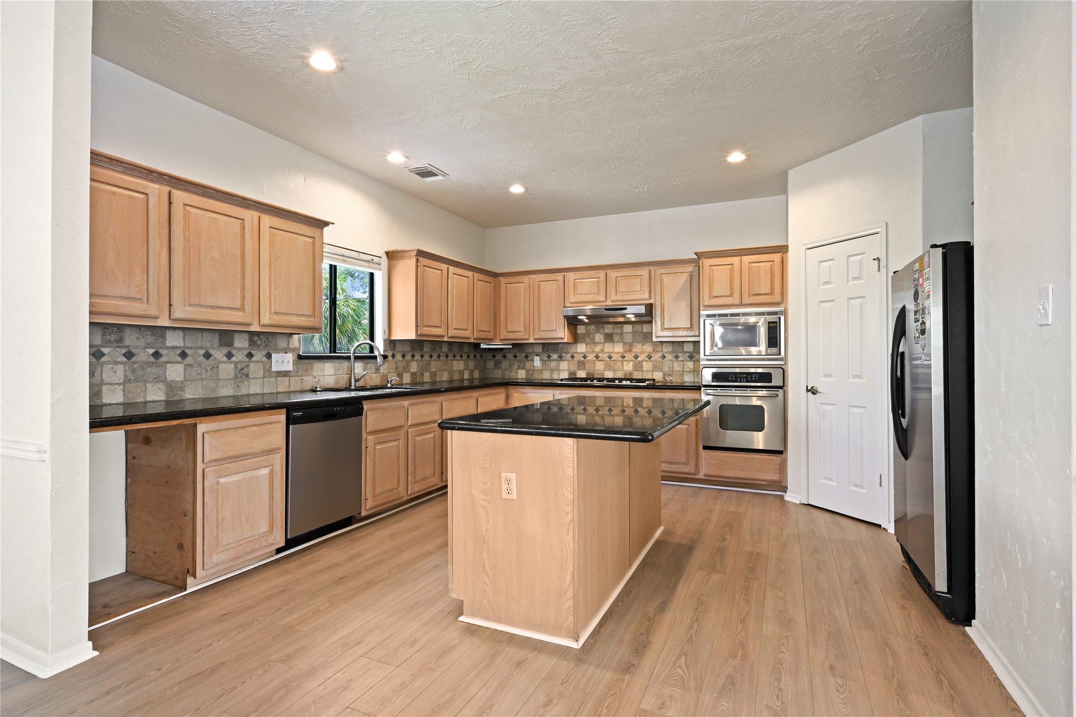 Kitchen accented with light wood cabinetry, sleek black countertops, and a central island, complemented by stainless steel appliances and a stylish tile backsplash.