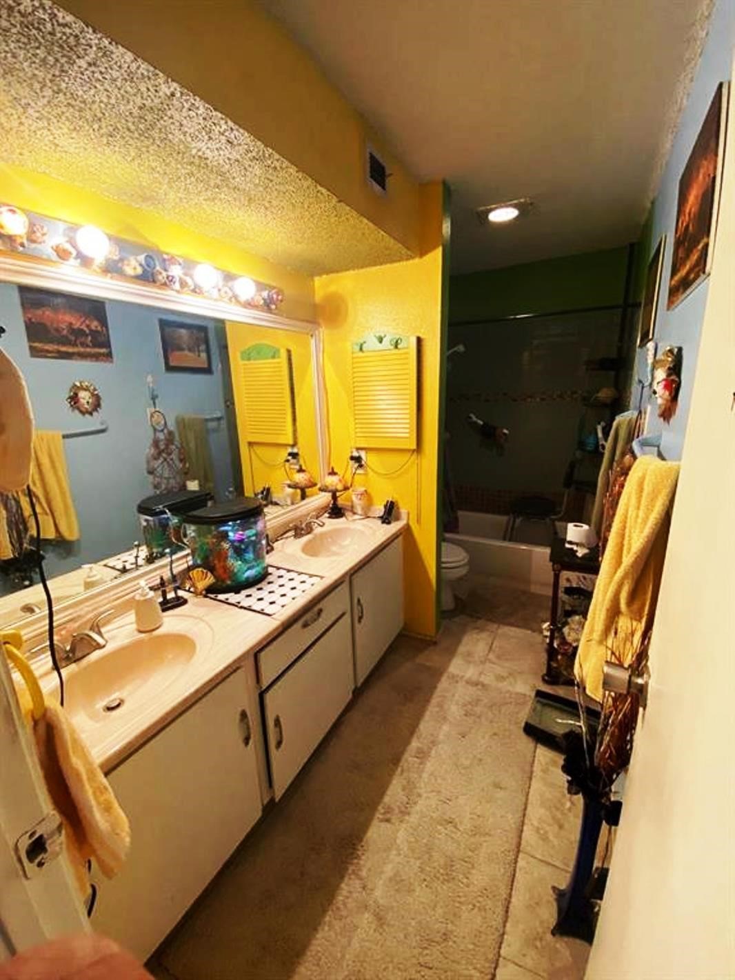 This bathroom features vibrant yellow and blue walls, dual sinks with a large mirror, and ample lighting. It includes a shower-tub combo and has quirky decorative elements, creating a unique and colorful atmosphere.