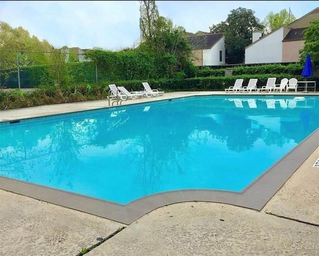 This photo features a spacious outdoor swimming pool surrounded by a concrete deck, with several lounge chairs for relaxation. It's set against a backdrop of greenery and nearby houses, offering a serene and inviting atmosphere for residents.
