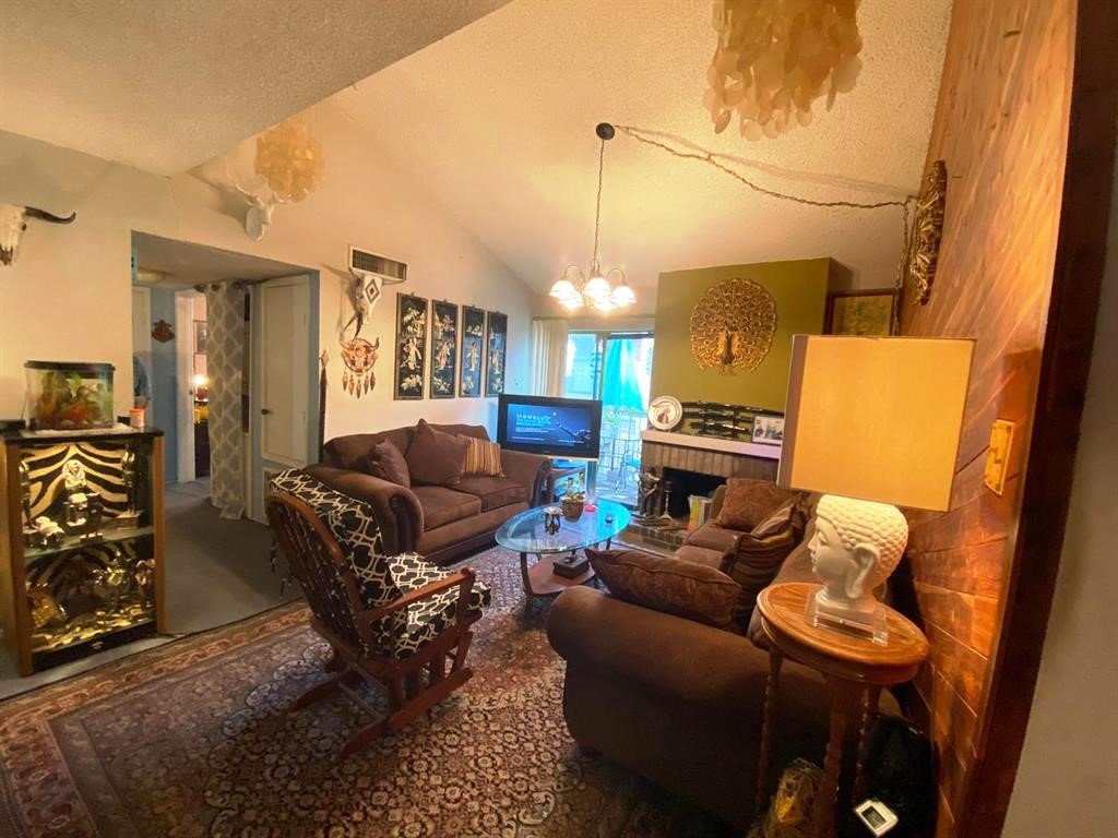 This cozy living room features vaulted ceilings, warm lighting, and a mix of eclectic decor. A comfortable seating area is centered around a glass coffee table, with a fireplace as a focal point. The space is accented with art pieces and a plush rug, offering a welcoming and unique atmosphere.
