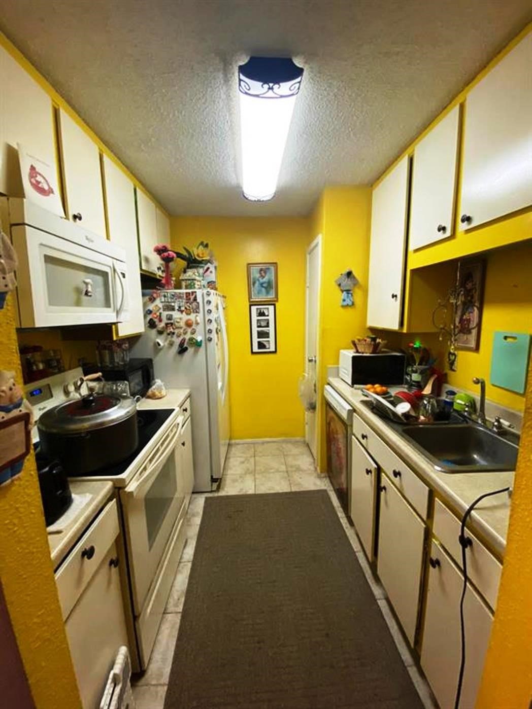 This cozy, narrow kitchen features bright yellow walls and white cabinetry, equipped with essential appliances like a stove, microwave, and refrigerator. It has overhead lighting, tile flooring, and ample storage space, creating a warm and functional cooking area.