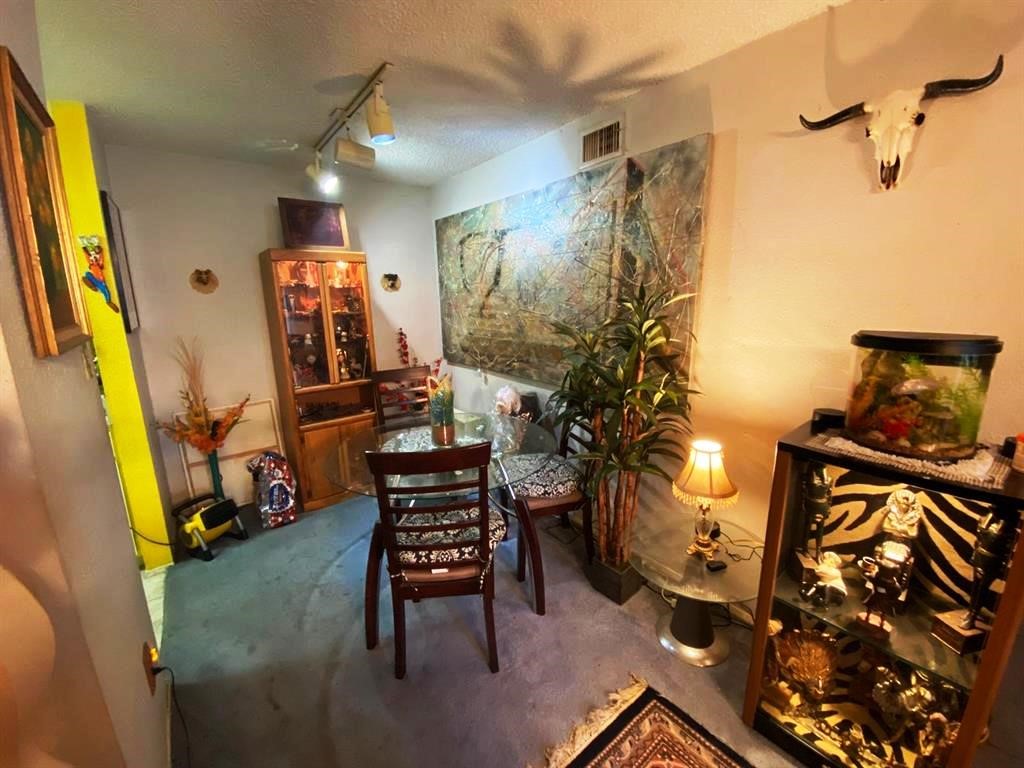 Cozy dining area with unique decor, featuring a glass table and chairs, art pieces, and display cabinets. The room has a warm ambiance with soft lighting and decorative items like a fish tank and wall-mounted skull.