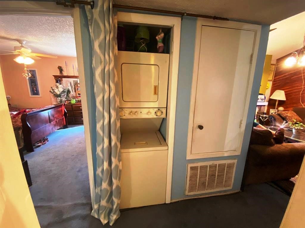 This photo shows a compact laundry area with a stacked washer and dryer unit, separated by a curtain. It is situated between a cozy living space and a bedroom, featuring a warm color palette and carpeted flooring.