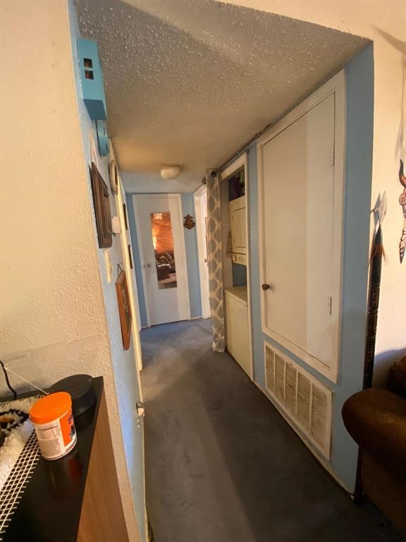 A narrow hallway with textured ceilings, leads to a room with a partially open door. It features a built-in cabinet, a small utility area with a stacked washer and dryer, and a carpeted floor.