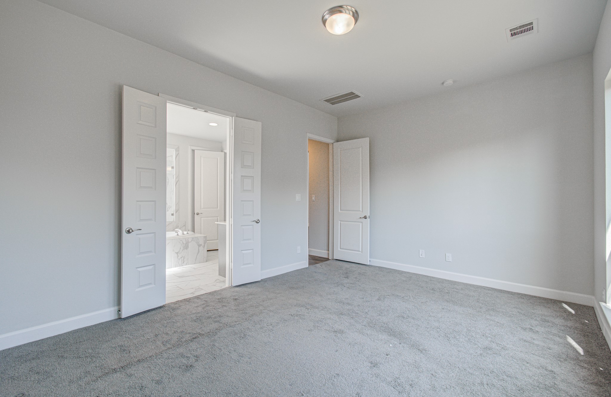 Spacious primary bedroom featuring soft carpeting, neutral walls, and ample natural light, with double doors opening to an elegant, tile-accented en-suite bathroom.