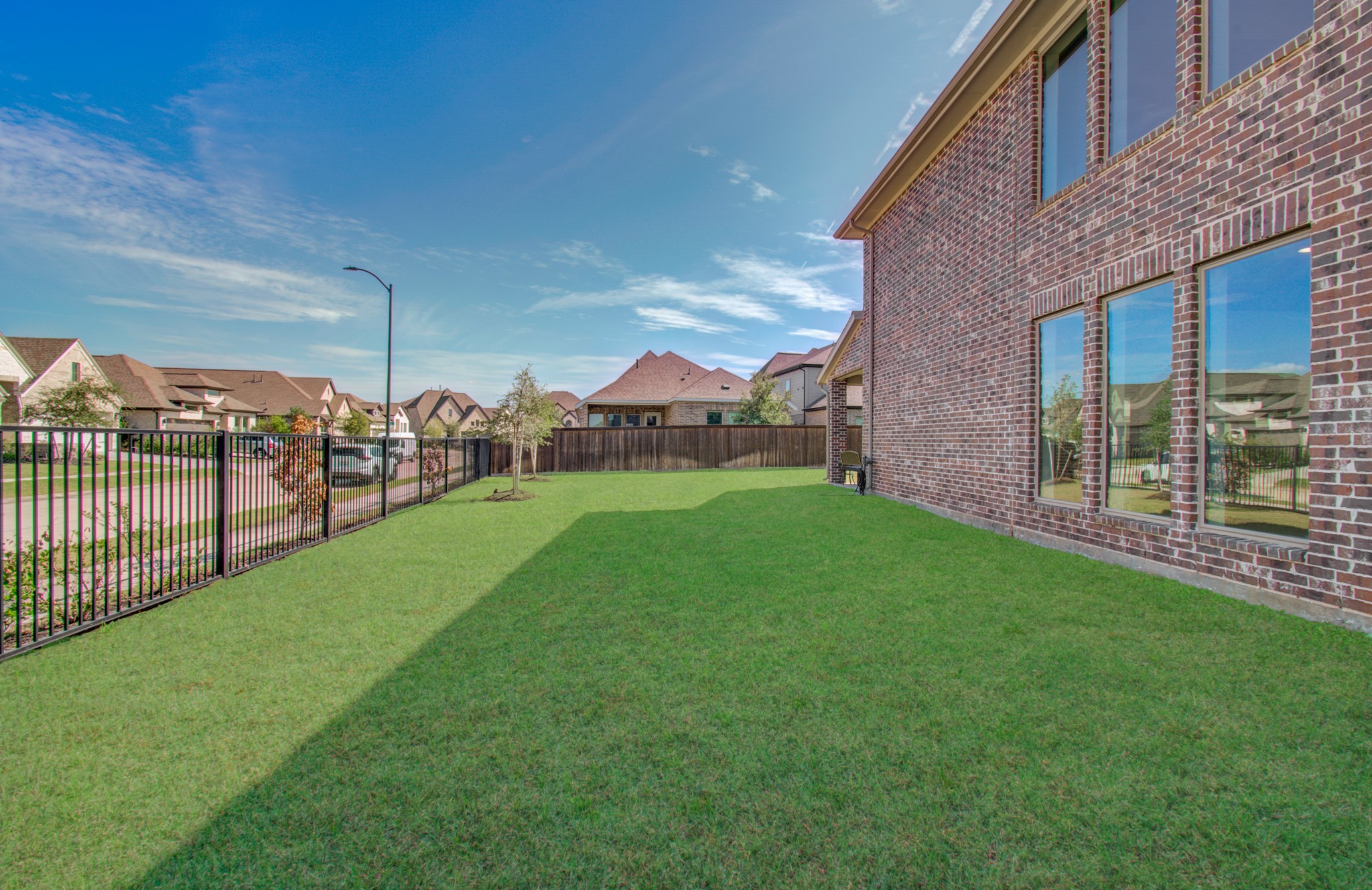 Spacious backyard with lush green lawn, bordered by a black metal fence, and large windows on a brick exterior offering ample natural light.