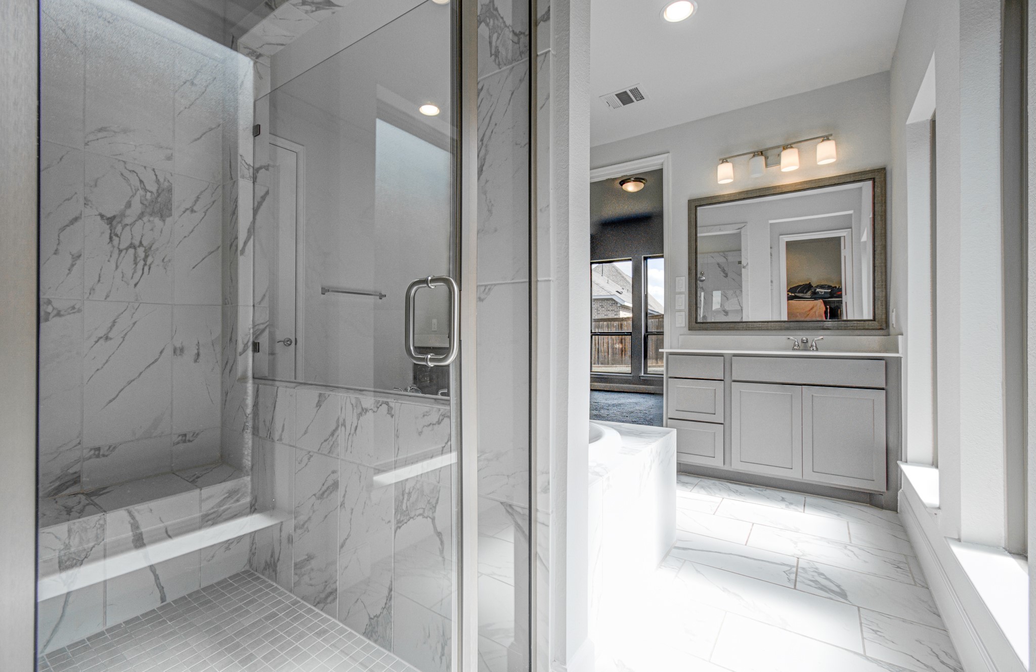 Elegant primary bathroom featuring a spacious glass-enclosed marble shower, modern vanity with framed mirror, and abundant natural light.