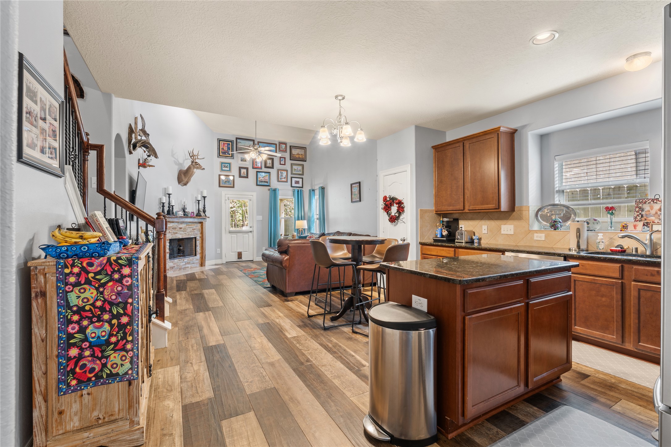 Spacious open-concept living area with warm wood flooring, a cozy stone fireplace, and a modern kitchen featuring granite countertops and rich cabinetry.