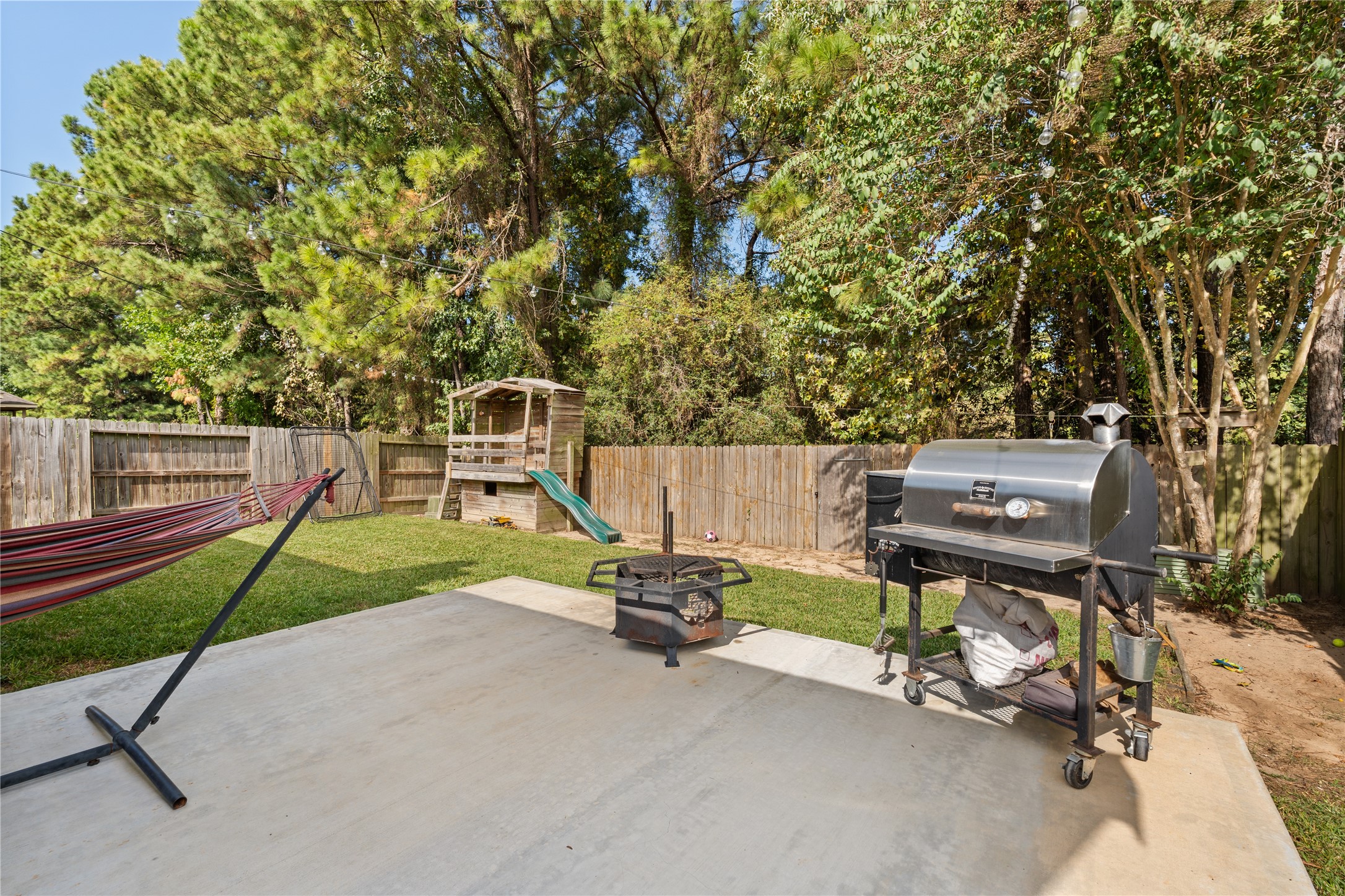 Spacious backyard with a concrete patio, perfect for grilling and relaxation, featuring a hammock and a play area with a slide, surrounded by lush greenery for privacy.