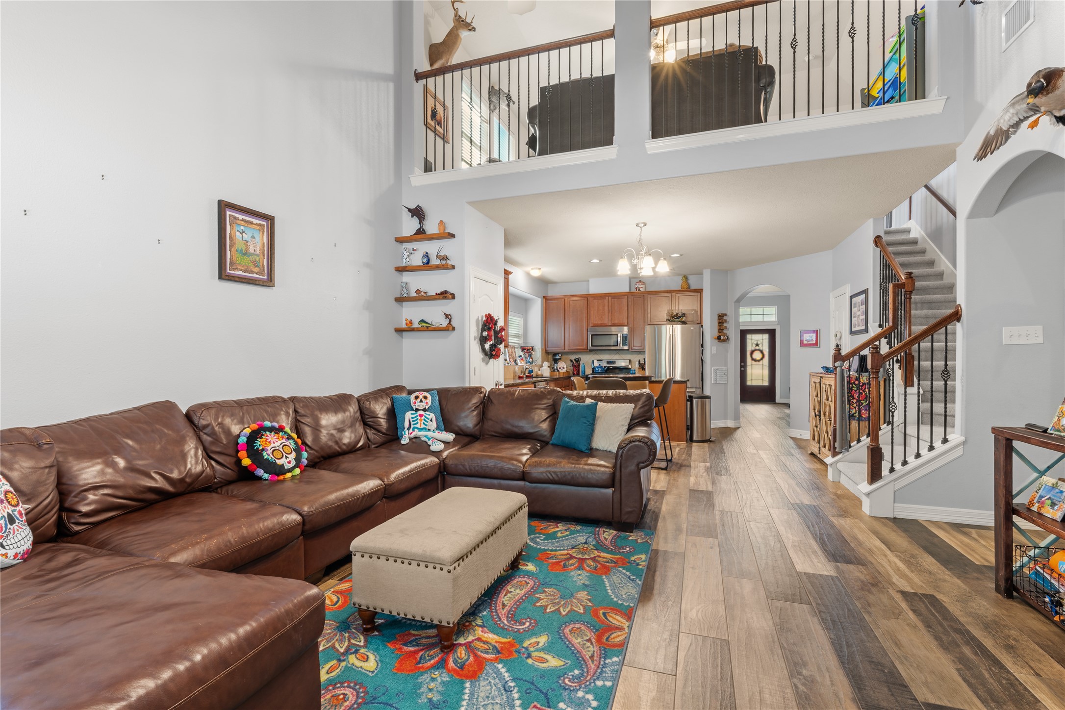 Spacious living area with high ceilings, featuring a cozy leather sectional and colorful decor. The open-concept design flows seamlessly into the kitchen, boasting modern appliances and warm wooden cabinetry. An inviting staircase leads to an upper loft, enhancing the home's airy, welcoming feel. Spacious living area with high ceilings, featuring a cozy leather sectional and colorful decor. The open-concept design flows seamlessly into the kitchen, boasting modern appliances and warm wooden cabinetry. An inviting staircase leads to an upper loft, enhancing the home's airy, welcoming feel.