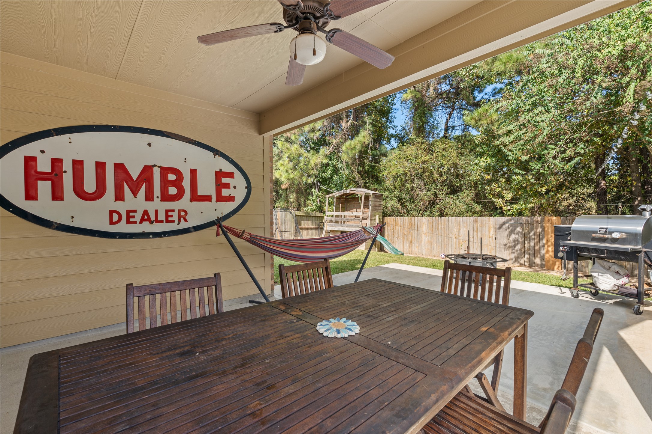 Charming covered patio with a ceiling fan, perfect for outdoor dining and relaxation, featuring a hammock, BBQ grill, and views of the lush backyard.