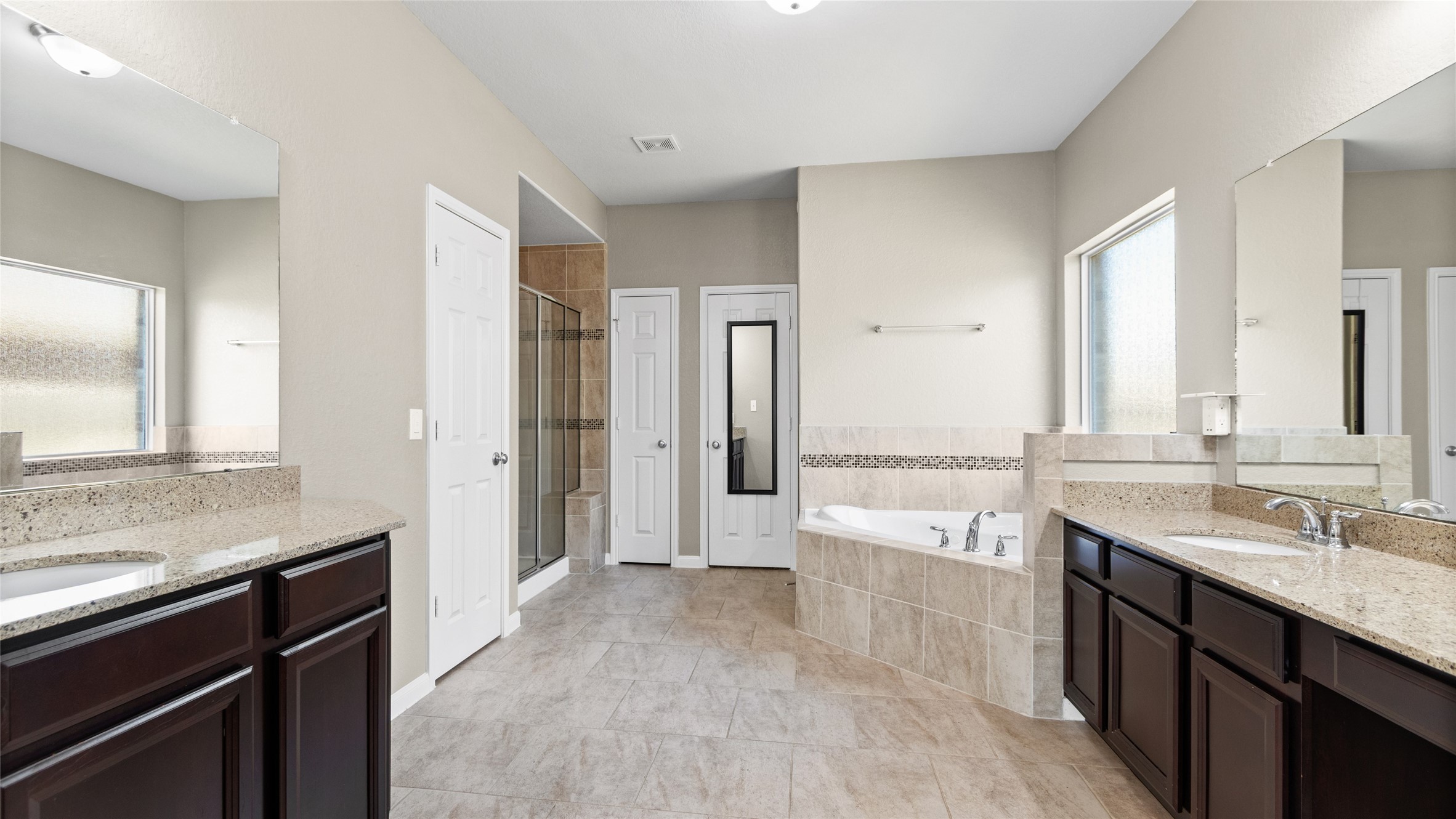 Primary suite bathroom featuring beautiful slate stone flooring, dual large vanities with ample storage, a deep jetted tub, a separate stone and glass shower, and a spacious walk-in closet.