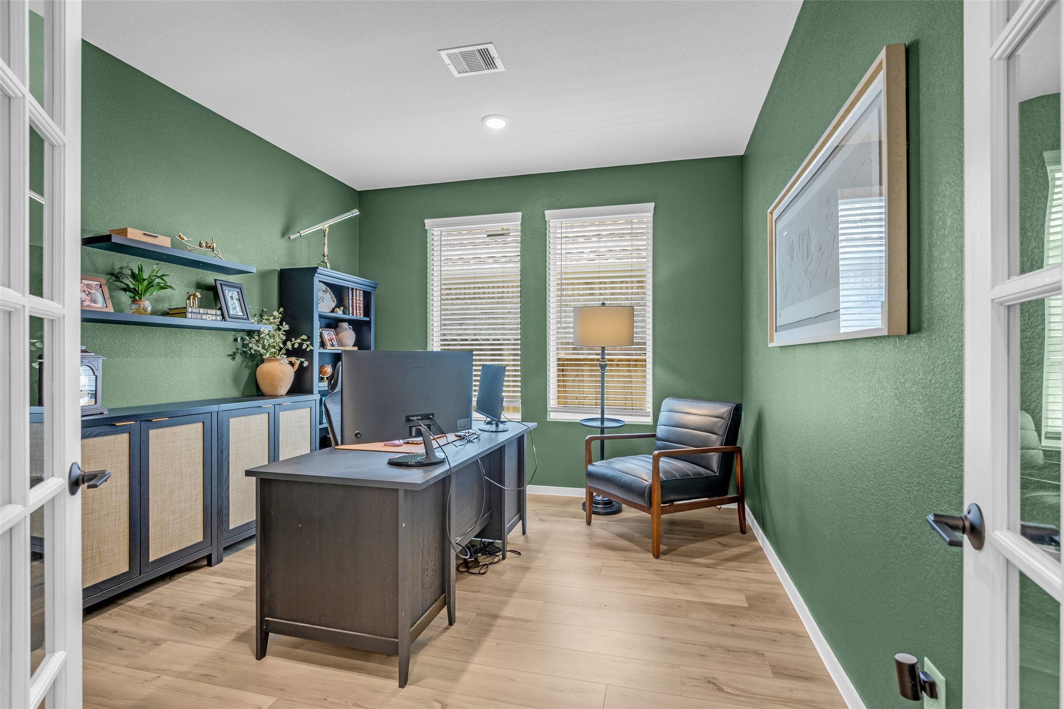 Elegant home office with rich green walls, featuring a sleek desk, cozy leather chair, ample natural light from two windows, and stylish built-in shelving for storage and decor.
