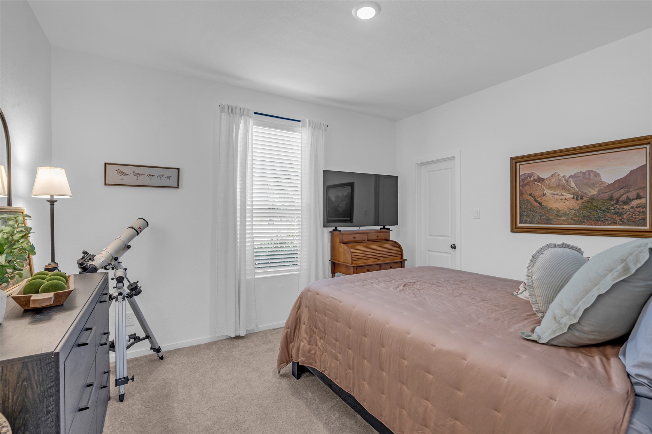 Cozy bedroom with natural light, featuring a comfortable bed, a telescope for stargazing, and tasteful decor including framed artwork.