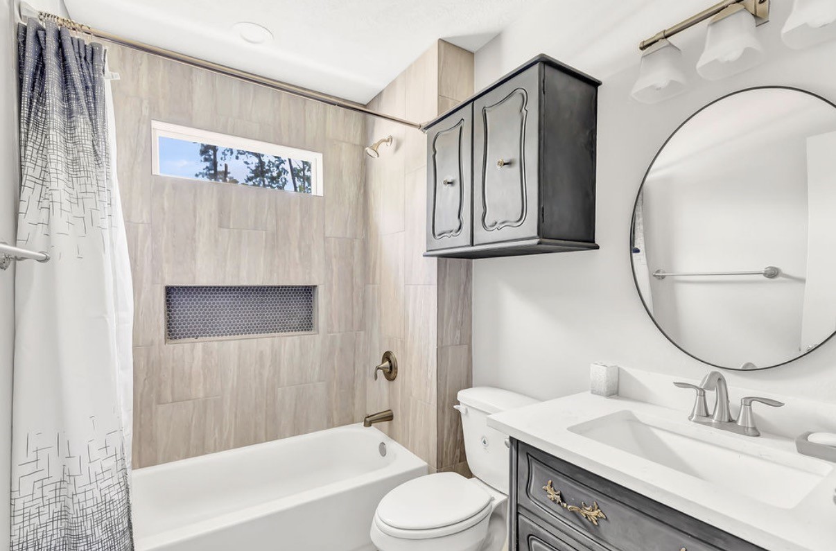 Experience modern elegance in this chic bathroom, featuring a sleek vanity with elegant fixtures, a stylish oval mirror, and a classic shower-tub combo framed by sophisticated tile work and a convenient built-in niche.