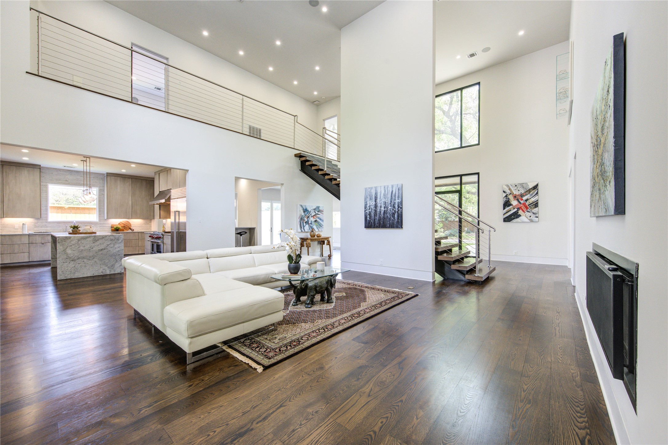 A spacious open-concept living area with high ceilings and abundant natural light. It includes a sleek kitchen with a large island, contemporary finishes, and a floating staircase leading to an upper level. The stylish design is complemented by dark wood floors, perfect for comfortable living and entertaining.