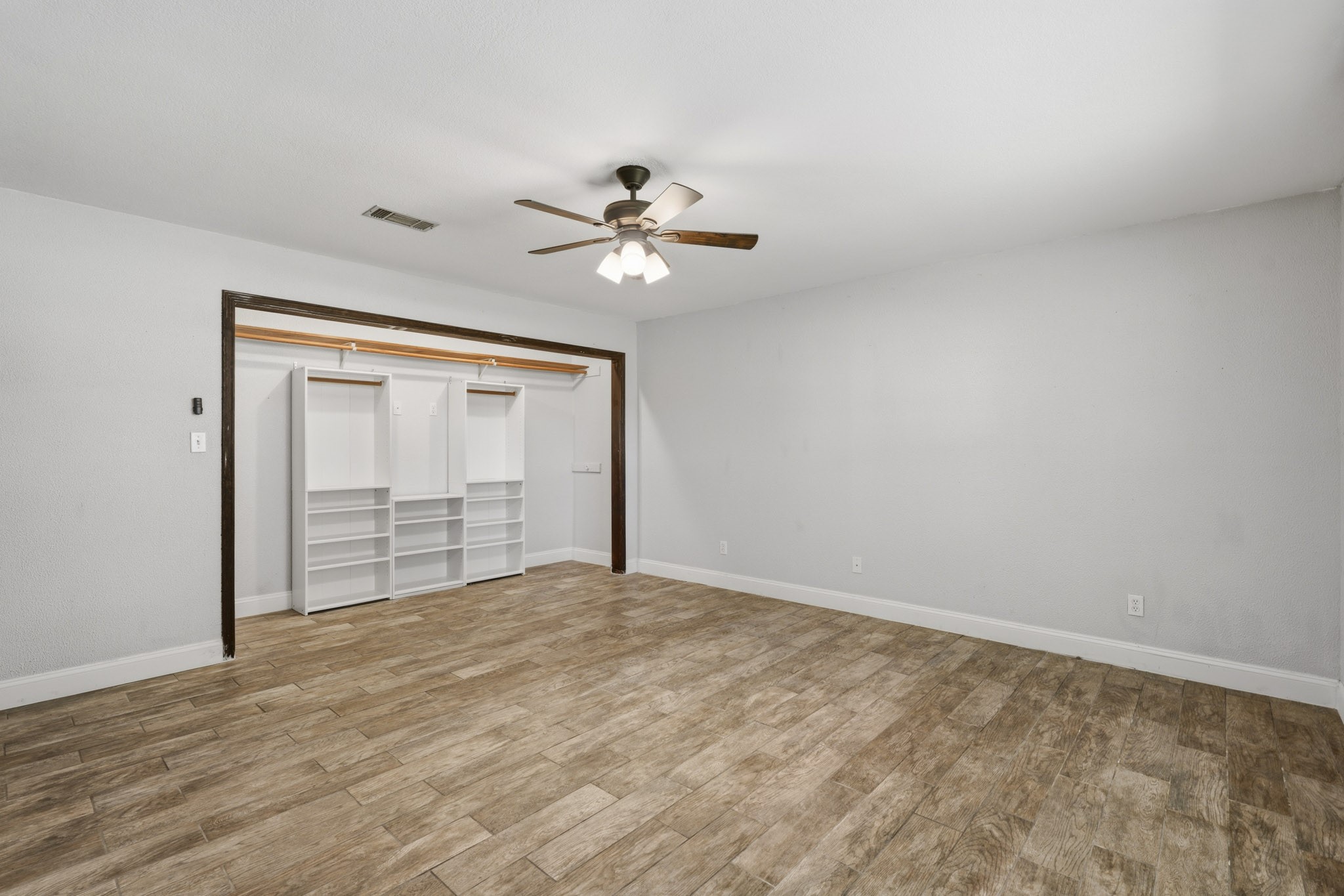 Spacious third bedroom with elegant wood-look tile flooring, featuring a ceiling fan and a large open closet with ample shelving for organized storage.