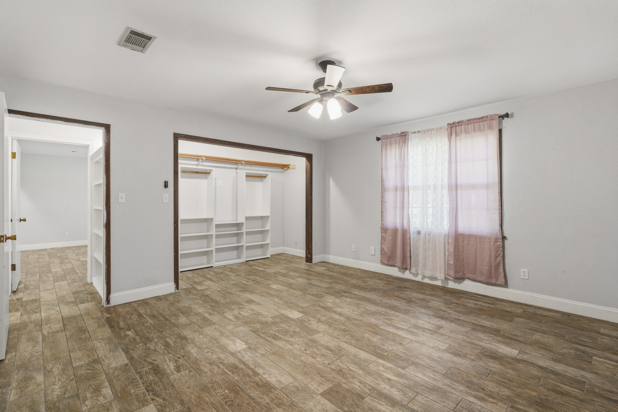 Spacious secondary bedroom featuring wood-look tile flooring, ample closet space with built-in shelving, and a ceiling fan for comfort. Natural light streams through the window, enhancing the room's warmth.
