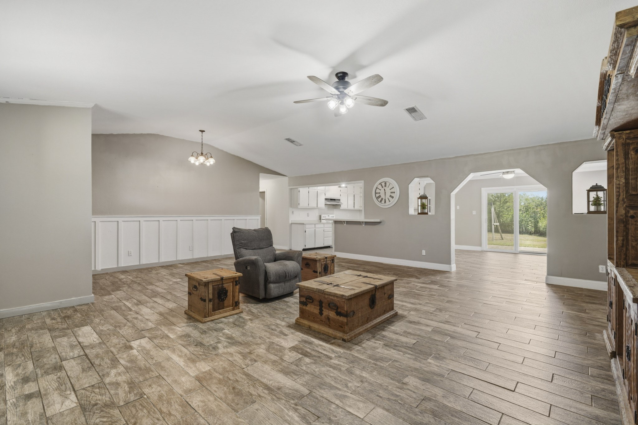 Spacious open-concept living area with wood-look tile flooring. The adjoining kitchen offers white cabinetry and modern lighting, leading to a bright dining area with sliding glass doors that open to a lush backyard view. Spacious open-concept living area with wood-look tile flooring. The adjoining kitchen offers white cabinetry and modern lighting, leading to a bright dining area with sliding glass doors that open to a lush backyard view.