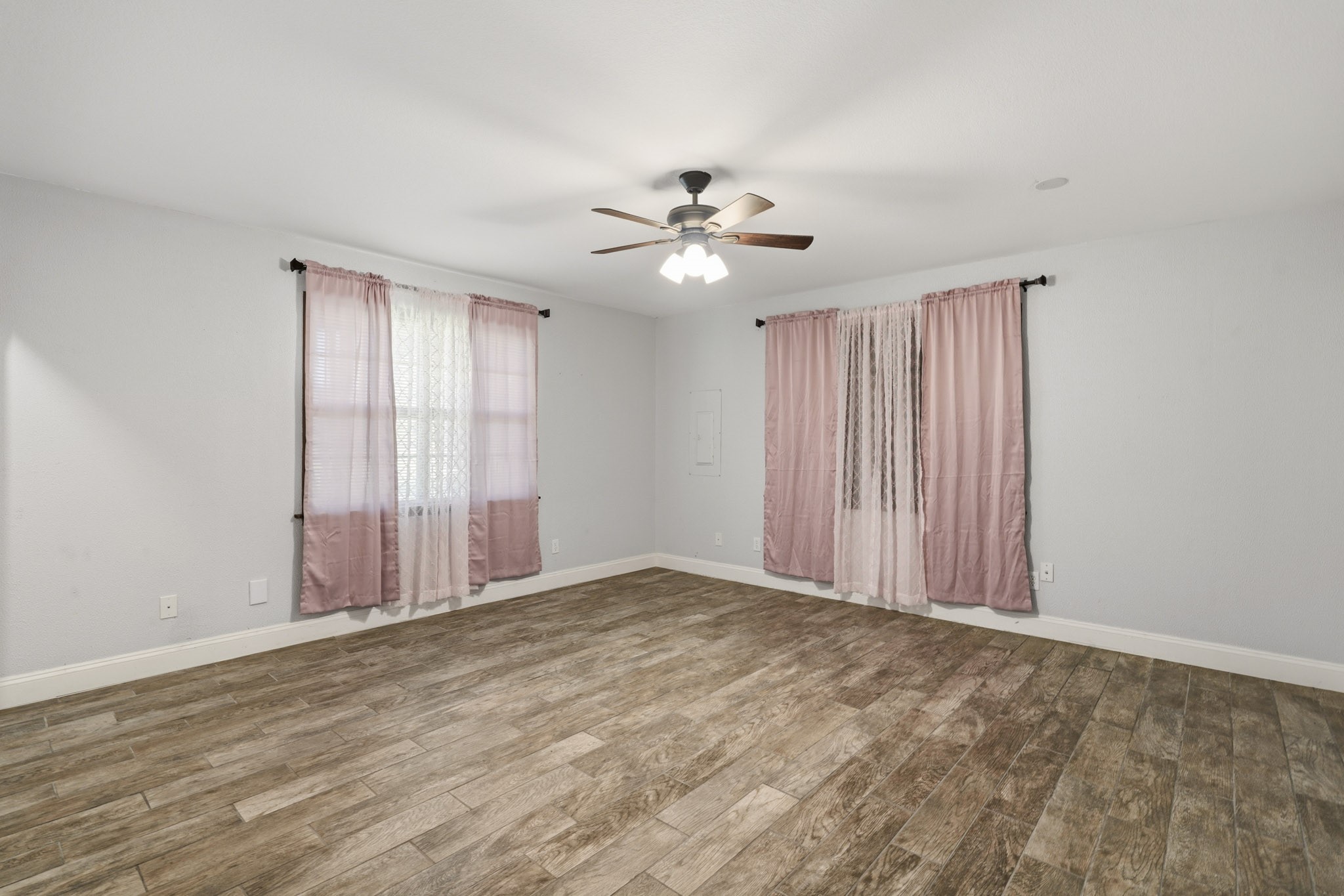 Spacious secondary room with warm wood-look flooring, soft gray walls, and ample natural light through large windows adorned with pink and white curtains. A ceiling fan adds a stylish touch.