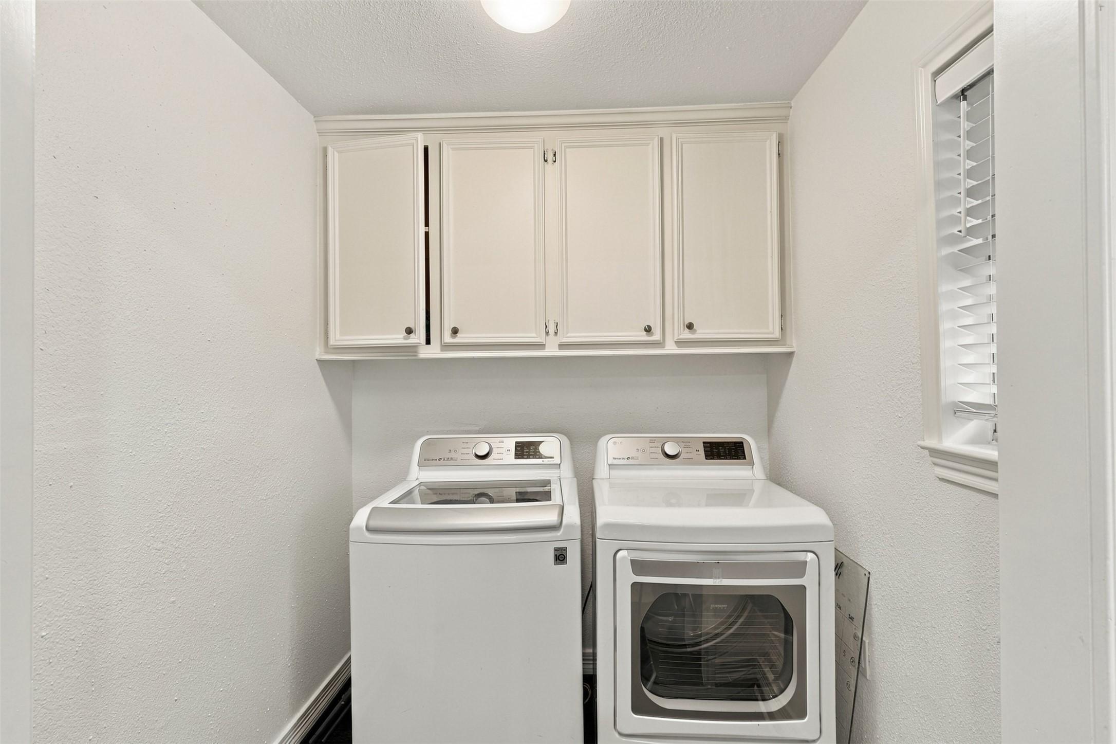 Bright and functional laundry room featuring modern washer and dryer, ample storage with overhead cabinetry, and a convenient window for natural light.