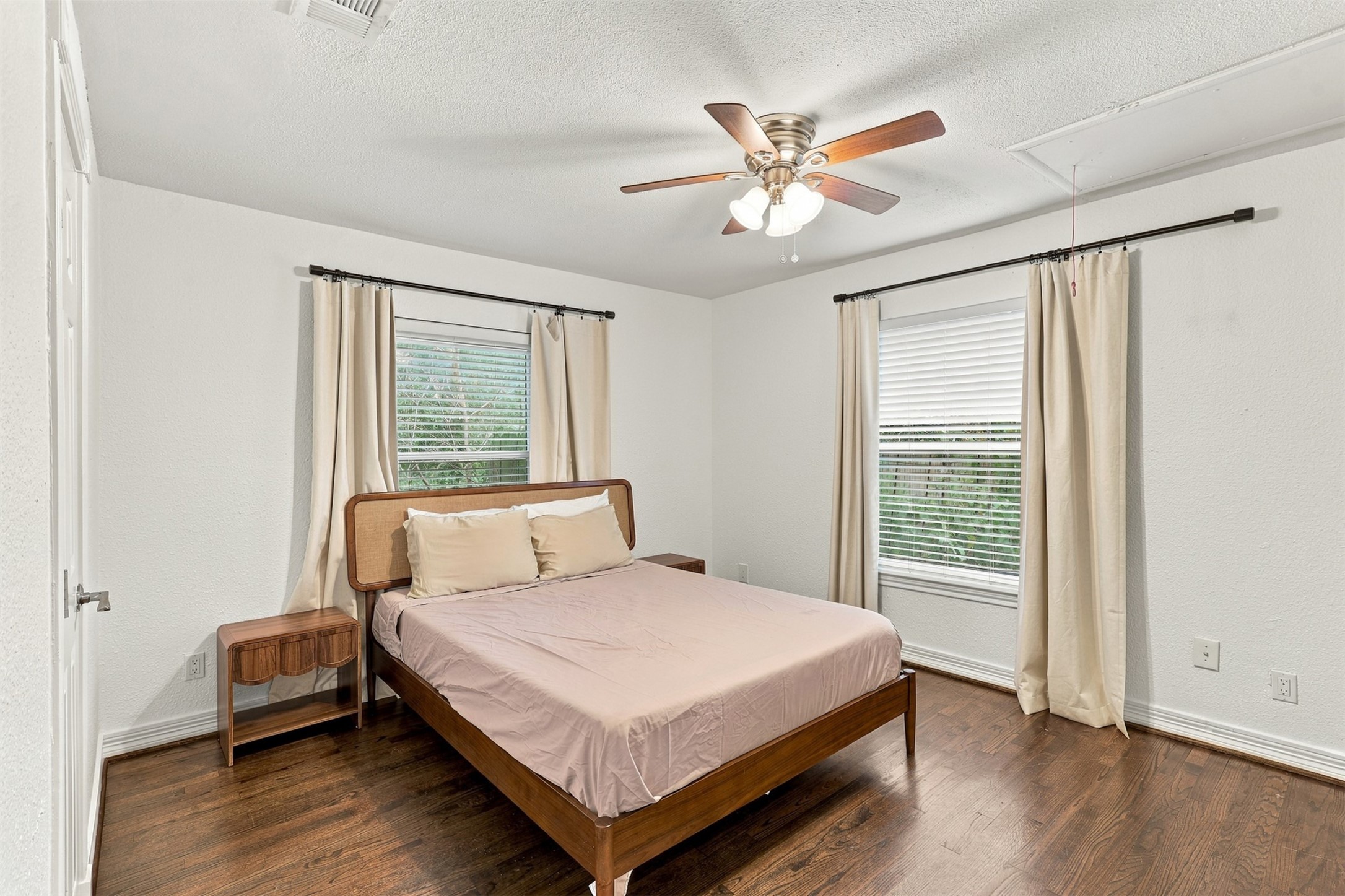 Bright and inviting bedroom with hardwood floors, featuring a comfortable bed, dual windows for natural light, and a ceiling fan for added comfort.