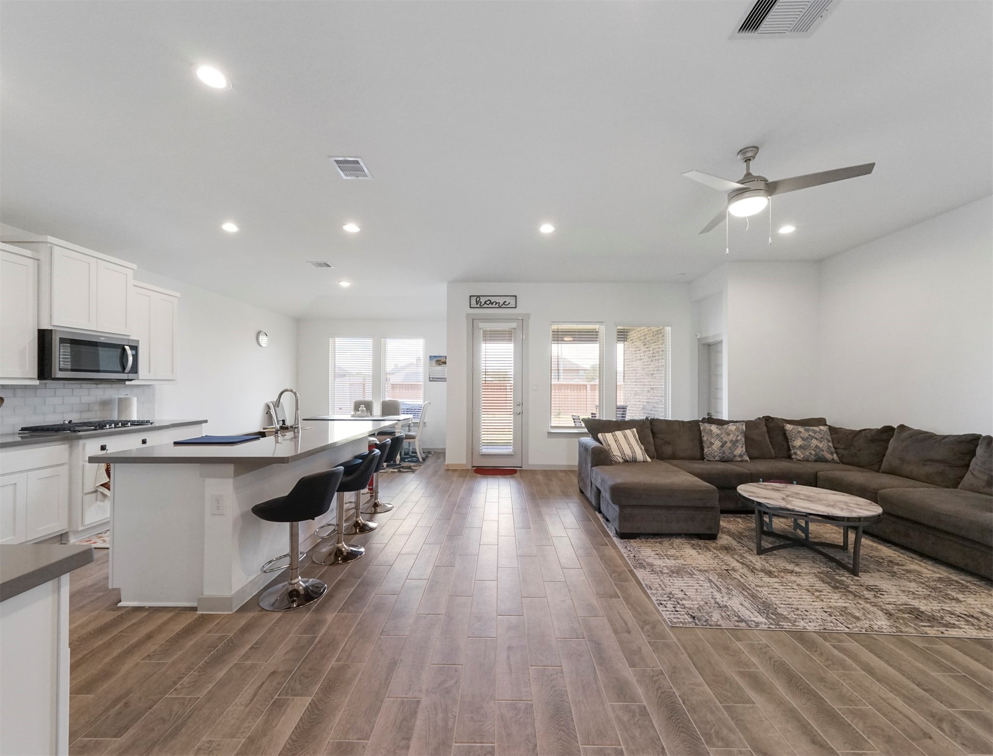 An open layout connects the kitchen, dining area, and living room. Wood-look tile flooring runs throughout, with recessed lighting and a rear door to the backyard.