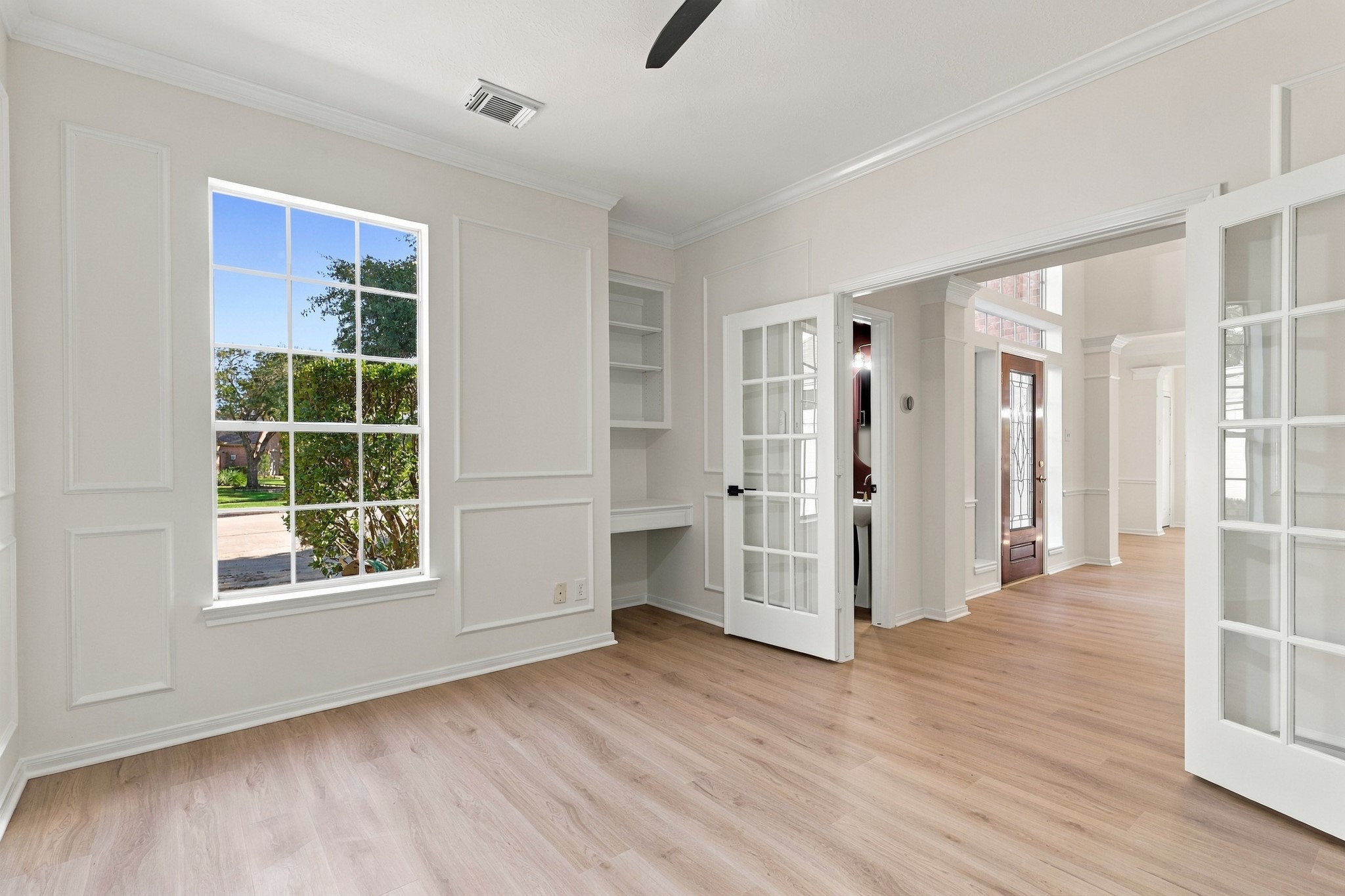 Bright and inviting home office with elegant French doors, built-in shelving, and abundant natural light from the large window, complementing the warm wood flooring.