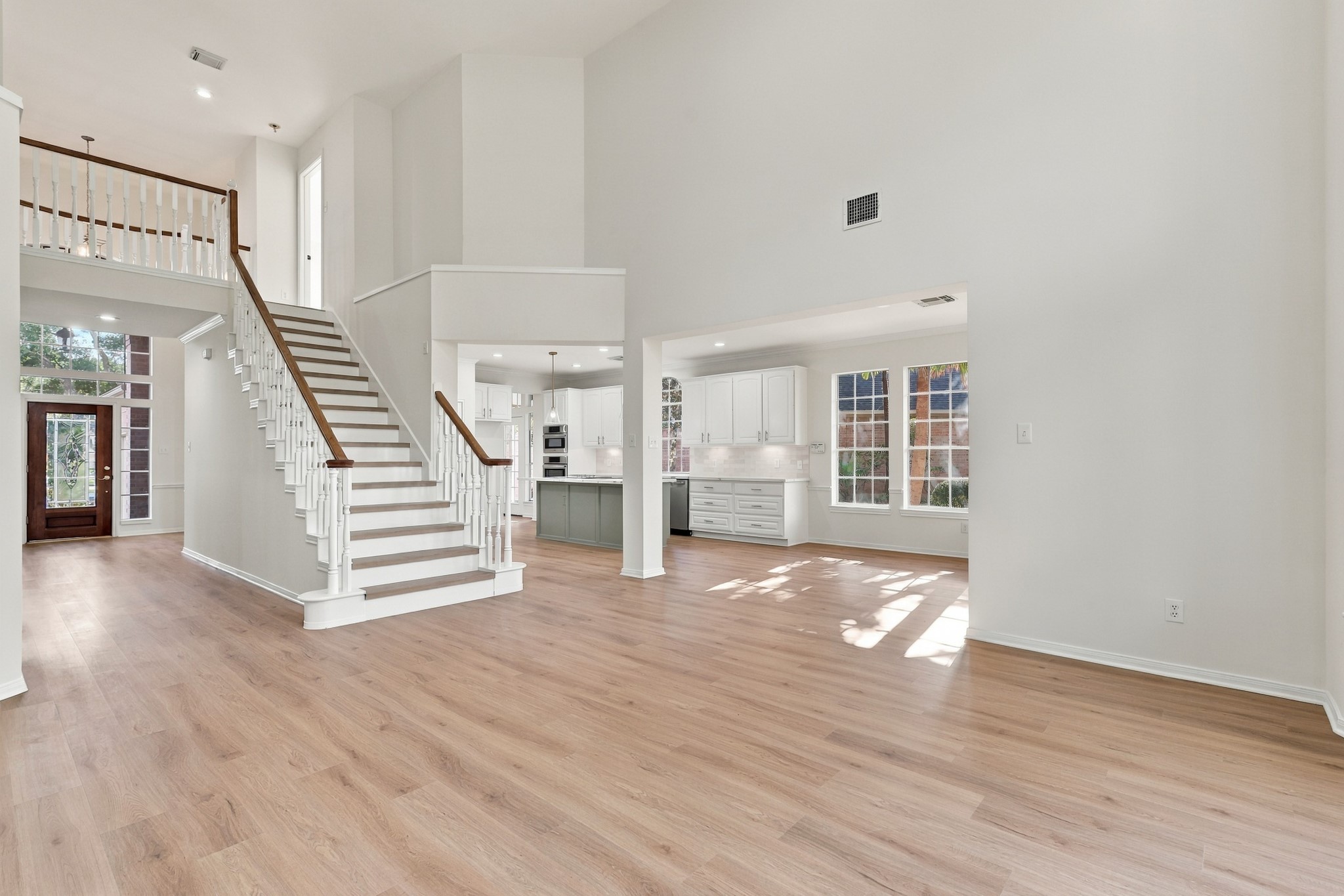 Spacious open-concept living area with high ceilings, featuring a grand staircase and abundant natural light from large windows, seamlessly flowing into a modern kitchen with sleek cabinetry.