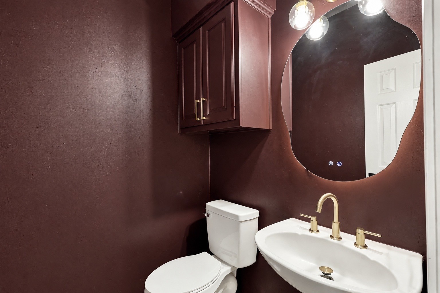 Chic powder room featuring rich burgundy walls, a modern round mirror, gold fixtures, and sleek cabinetry for a touch of luxury.