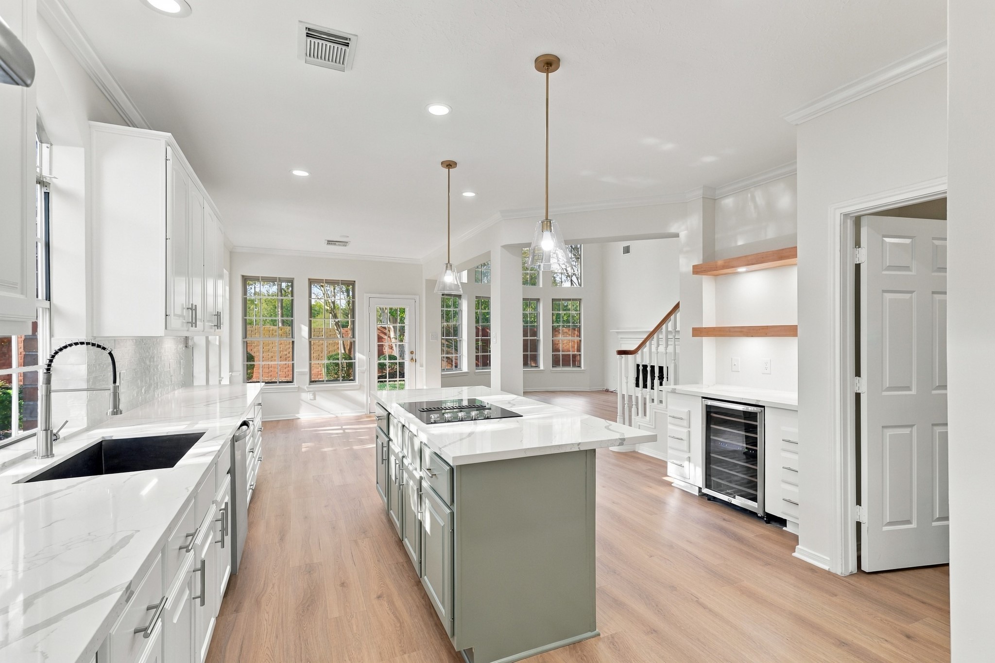 Bright and spacious open-concept kitchen featuring elegant white cabinetry, a large island with a cooktop, and sleek pendant lighting, seamlessly flowing into a sunlit living area with large windows and hardwood floors.
