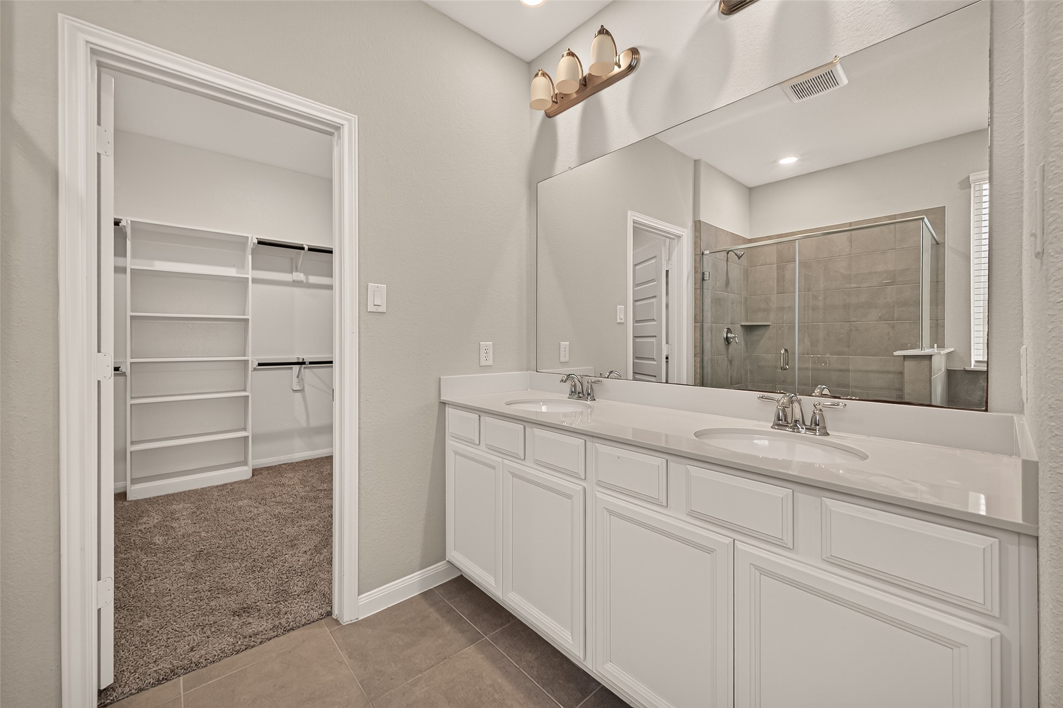 Elegant bathroom featuring a spacious double vanity, large mirror, and modern light fixtures, leading to a roomy walk-in closet and a sleek, glass-enclosed shower.