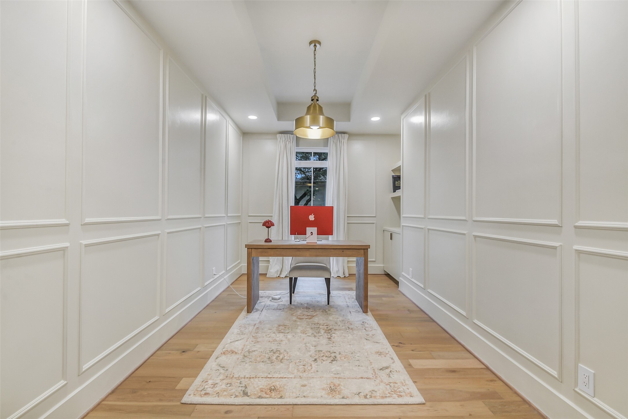 Millwork paneling covers every wall in this refined home office, adding architectural detail and a tailored, sophisticated feel. A statement brass pendant light hangs above the desk, with a sunlit window and built-in shelving completing the space.