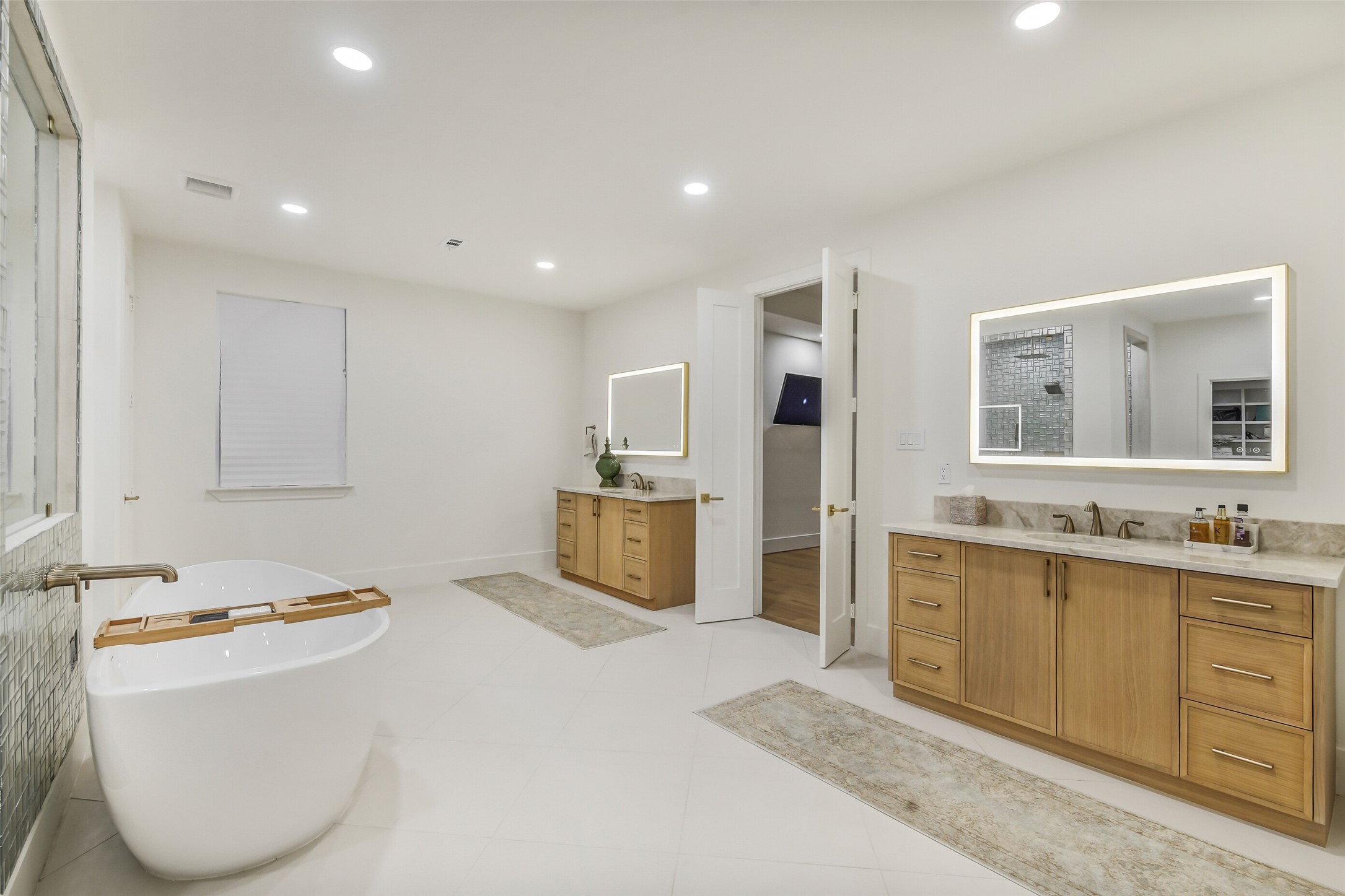 A spa-like primary bath featuring a freestanding soaking tub, two separate vanities with backlit mirrors, and a walk-in tiled shower. Warm wood cabinetry, quartz countertops, and thoughtful details throughout create a luxurious, private retreat.