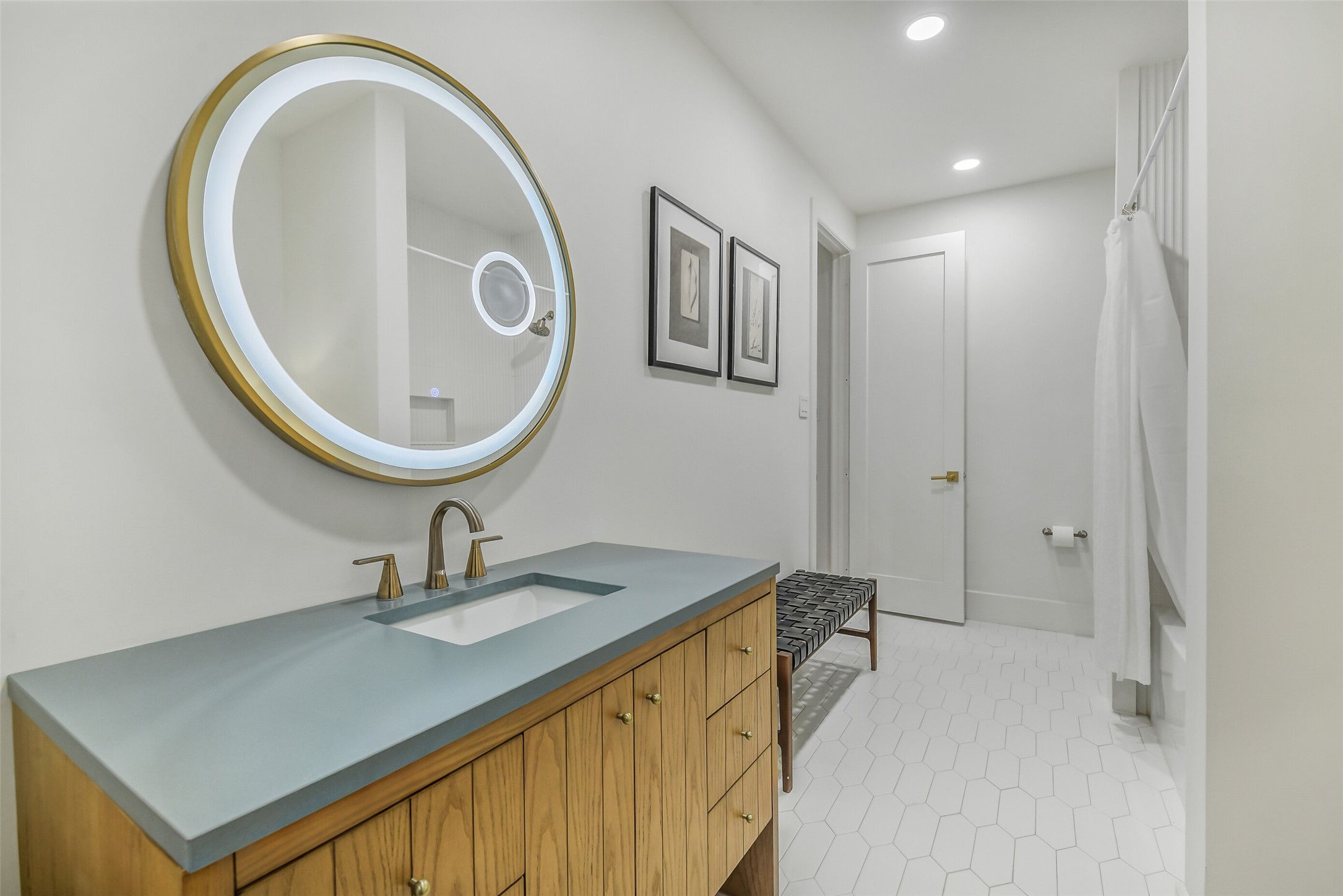 Gorgeous bath featuring a striking round backlit mirror in a brass frame, a blue concrete-look countertop, and warm wood cabinetry with brass hardware. Hexagon tile flooring and a woven bench add character throughout.