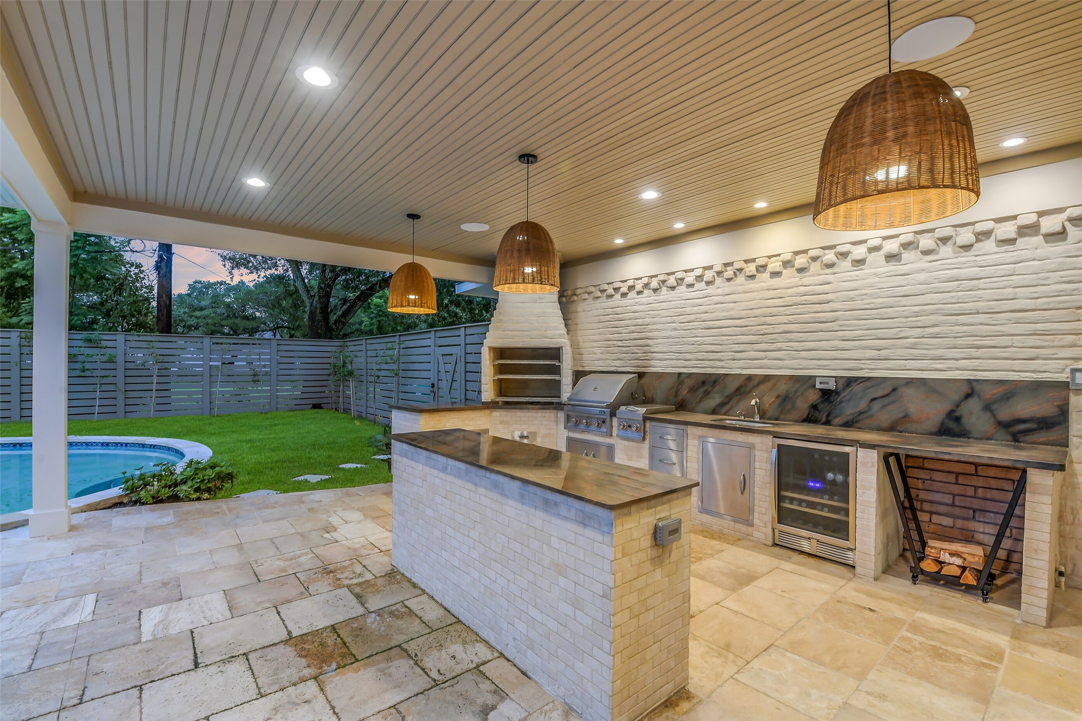 A true outdoor entertainer's dream, this fully equipped kitchen features a built-in grill, wine fridge, sink, and generous prep counters, all set beneath a woven pendant-lit wood ceiling. A brick pizza oven and wood-storage nook add rustic charm, while the space overlooks the pool and privately fenced backyard.