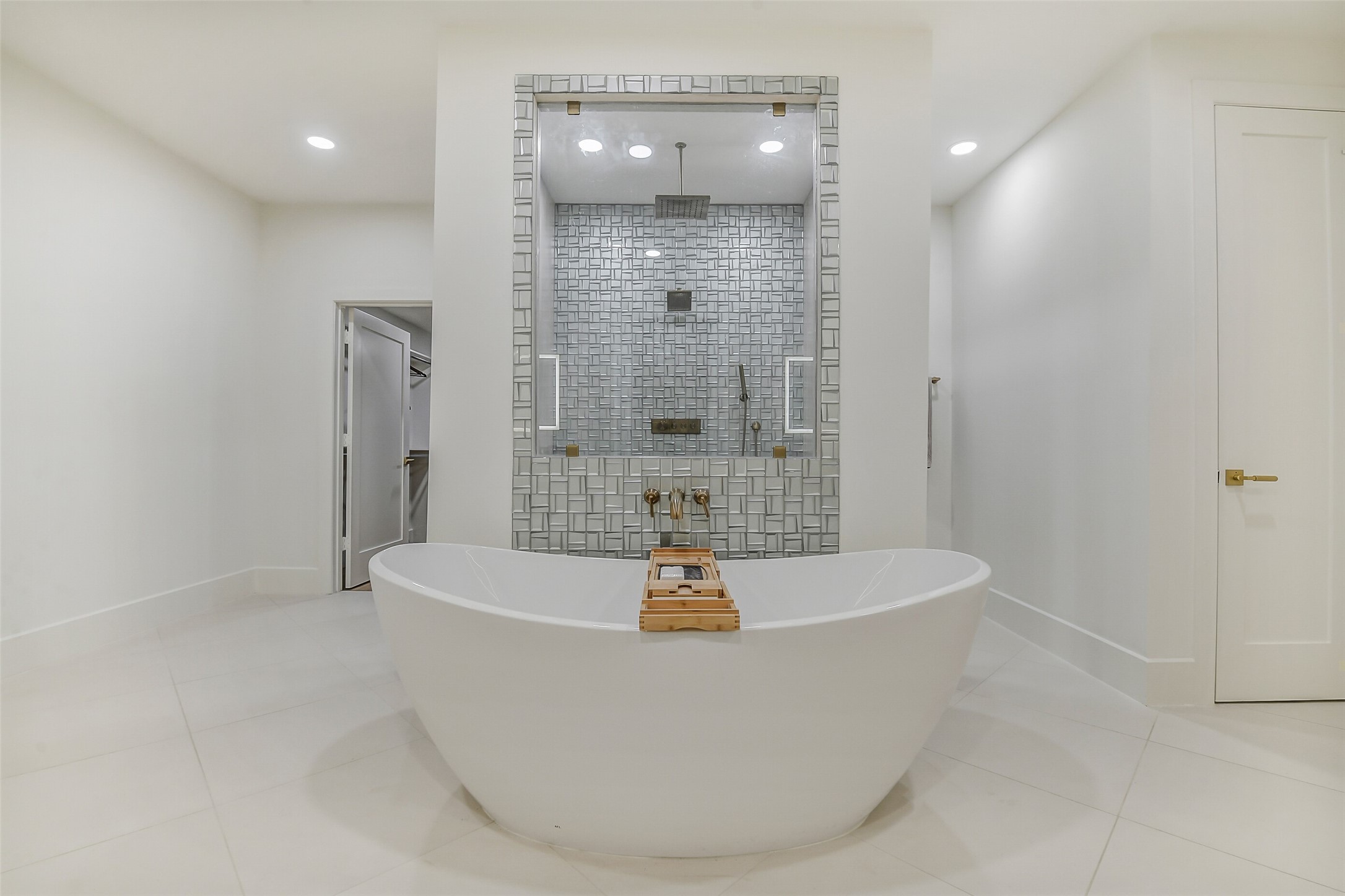 An oversized soaking tub sits front and center, framed by a stunning textured tile shower with a rain showerhead and handheld sprayer. Sleek brass fixtures and a spa-like walk-in closet nearby complete this serene, luxurious retreat.
