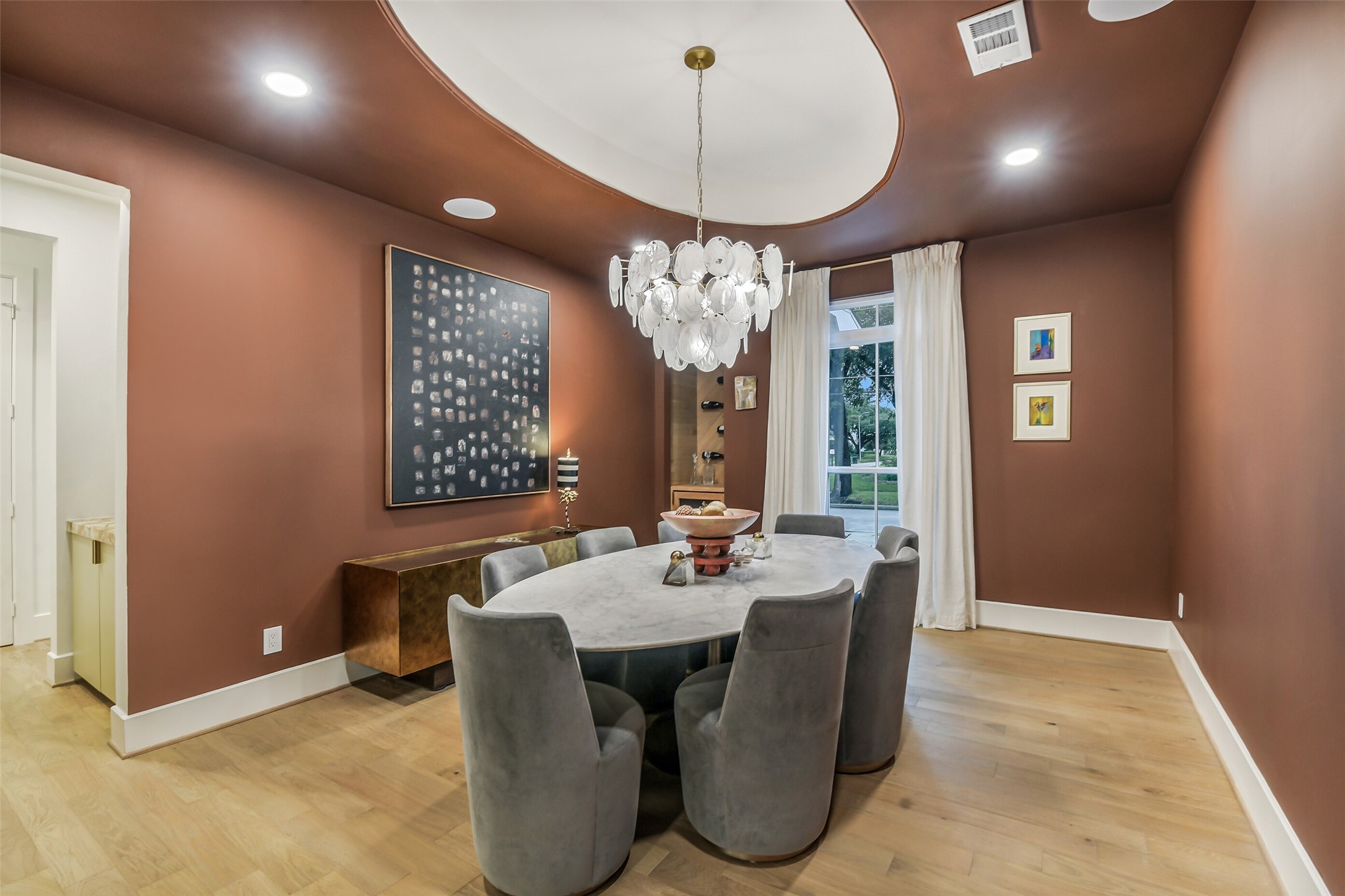 A rich, moody color palette sets the tone in this elegant dining room, anchored by a round table with plush swivel chairs beneath a sculptural bubble chandelier. A domed ceiling detail and curated artwork add a refined, sophisticated touch.