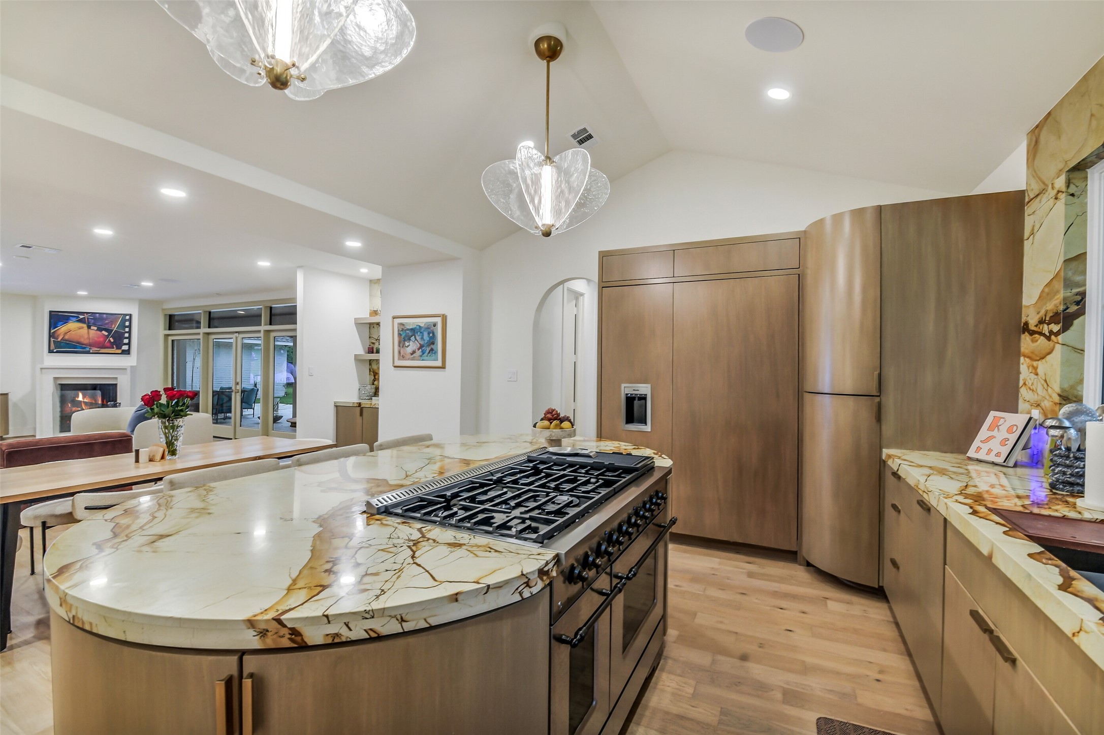 A show-stopping curved island anchors this chef's kitchen, wrapped in dramatic natural stone and fitted with a professional-grade gas range. Custom wood cabinetry conceals the built-in refrigerator, while sculptural glass pendants and a vaulted ceiling add elegance overhead.