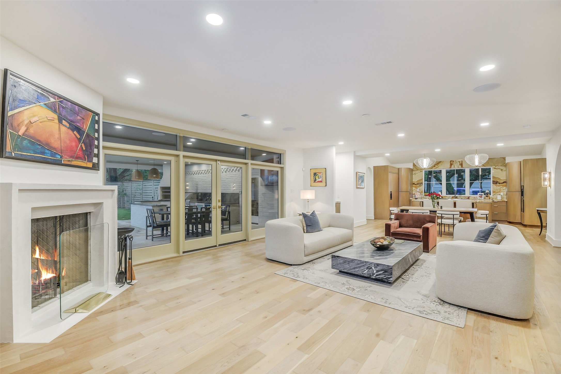 Bright, open living room centered around a sleep gas fireplace with a modern laster surround. Multi-panel glass doors lead out to the covered patio and outdoor kitchen, while the space flows seamlessly into the kitchen and dining areas beyond. Light wide-plank flooring, high ceilings and neutral pallet make this the ideal spot to relax or entertain.