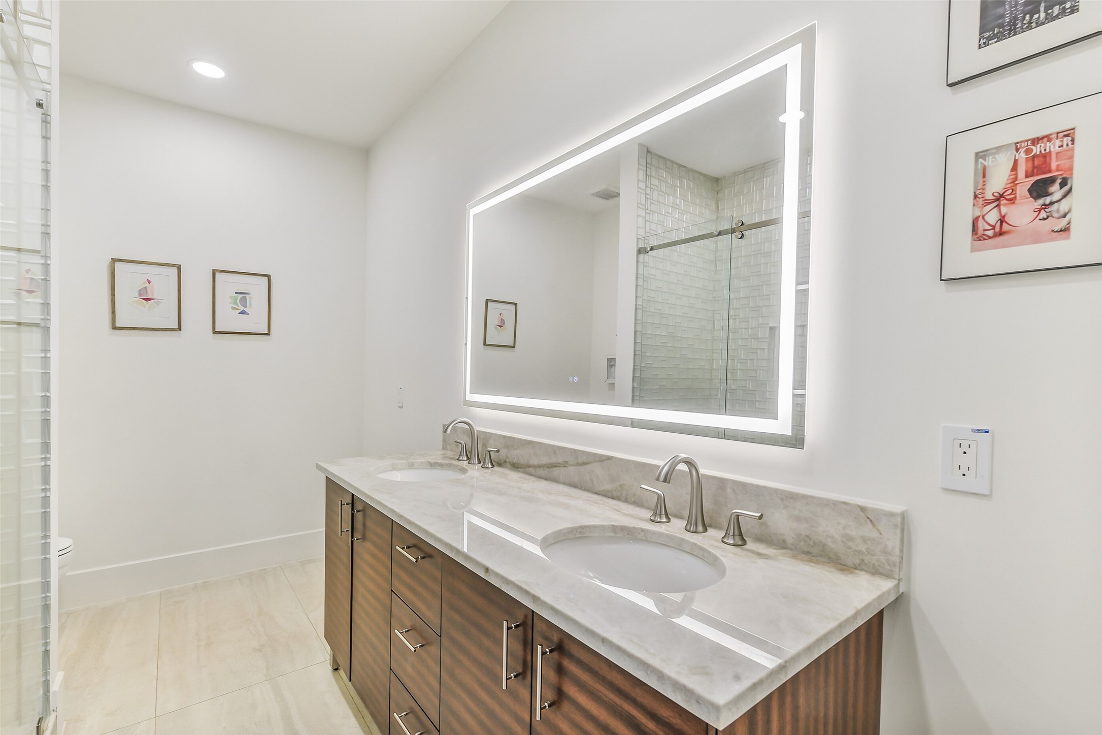 A sleek en-suite featuring a dual-sink vanity with rich wood cabinetry, a backlit LED mirror, and a glass-enclosed tiled shower. Stone countertops and modern fixtures give this bath a polished, upscale feel.