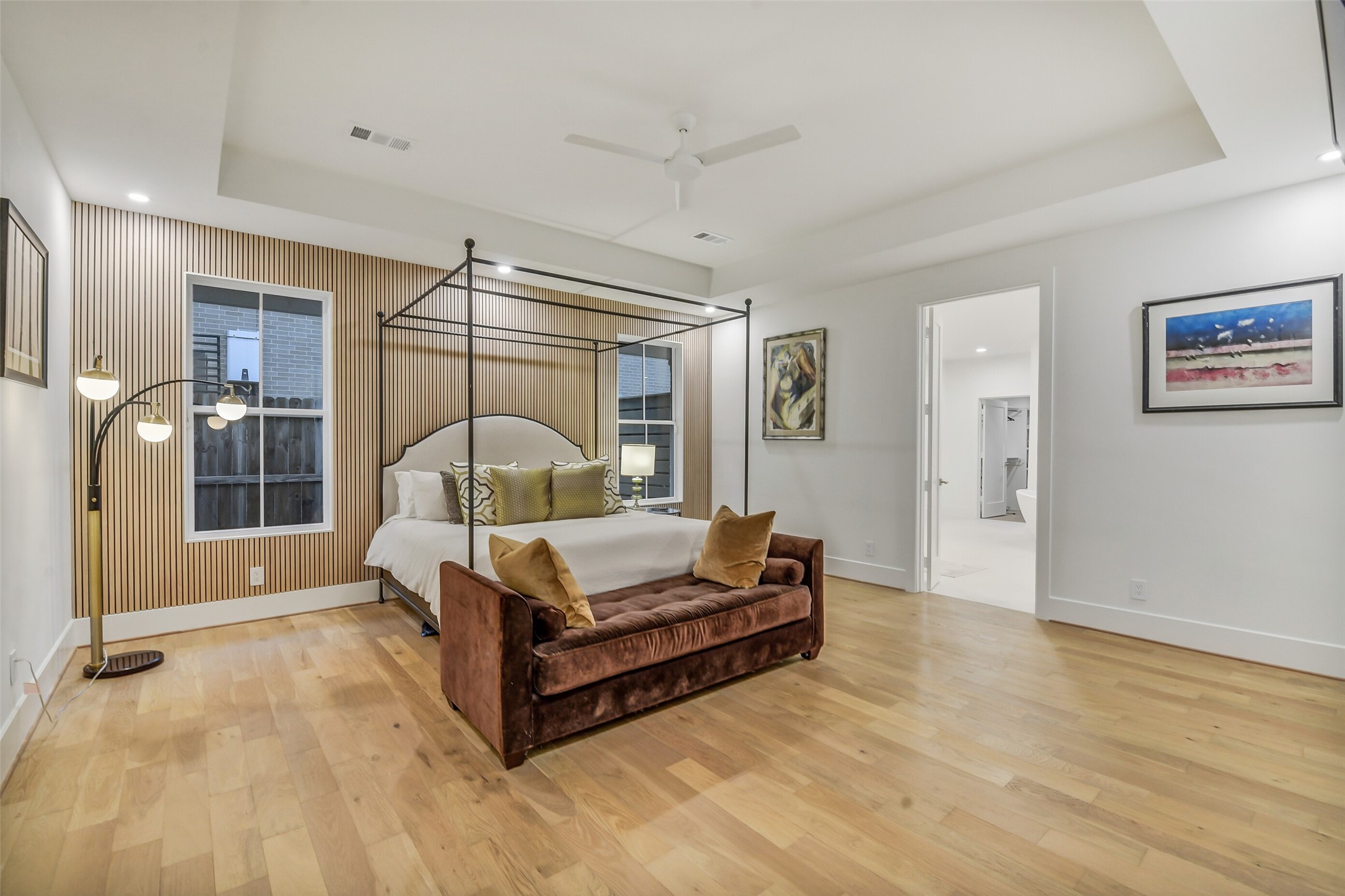 A spacious primary suite featuring a slatted wood accent wall behind the bed and a tray ceiling that adds architectural depth. A sleek canopy bed frame, plush velvet bench, and large windows create a warm, sophisticated retreat, with direct access to the en-suite bath just beyond.