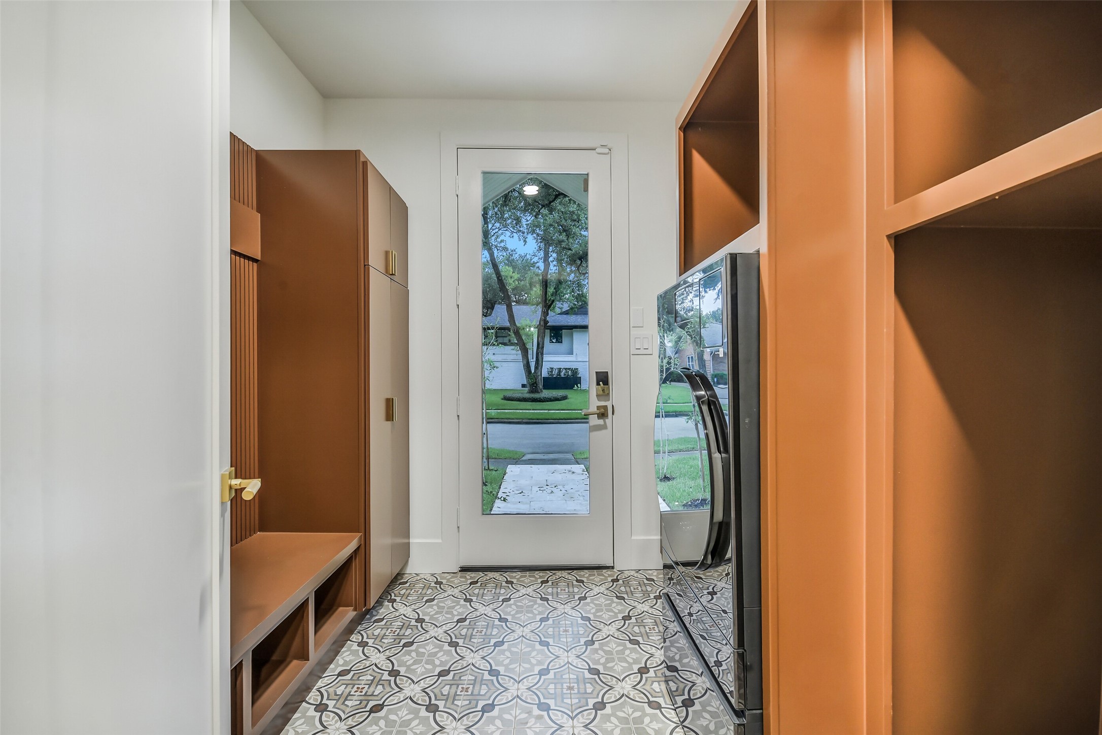 A stylish mudroom finished with patterned tile flooring and rich, custom cabinetry featuring fluted detailing and brass hardware. Built-in storage and a bench offer a functional drop zone, while a sleek all-in-one washer/dryer combo adds everyday convenience just steps from the back door.