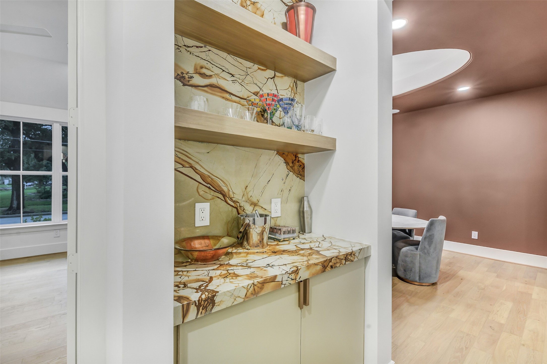 Tucked between the kitchen and dining room, this chic dry bar features open wood shelving and a striking stone slab backsplash, offering both storage and style for effortless entertaining.