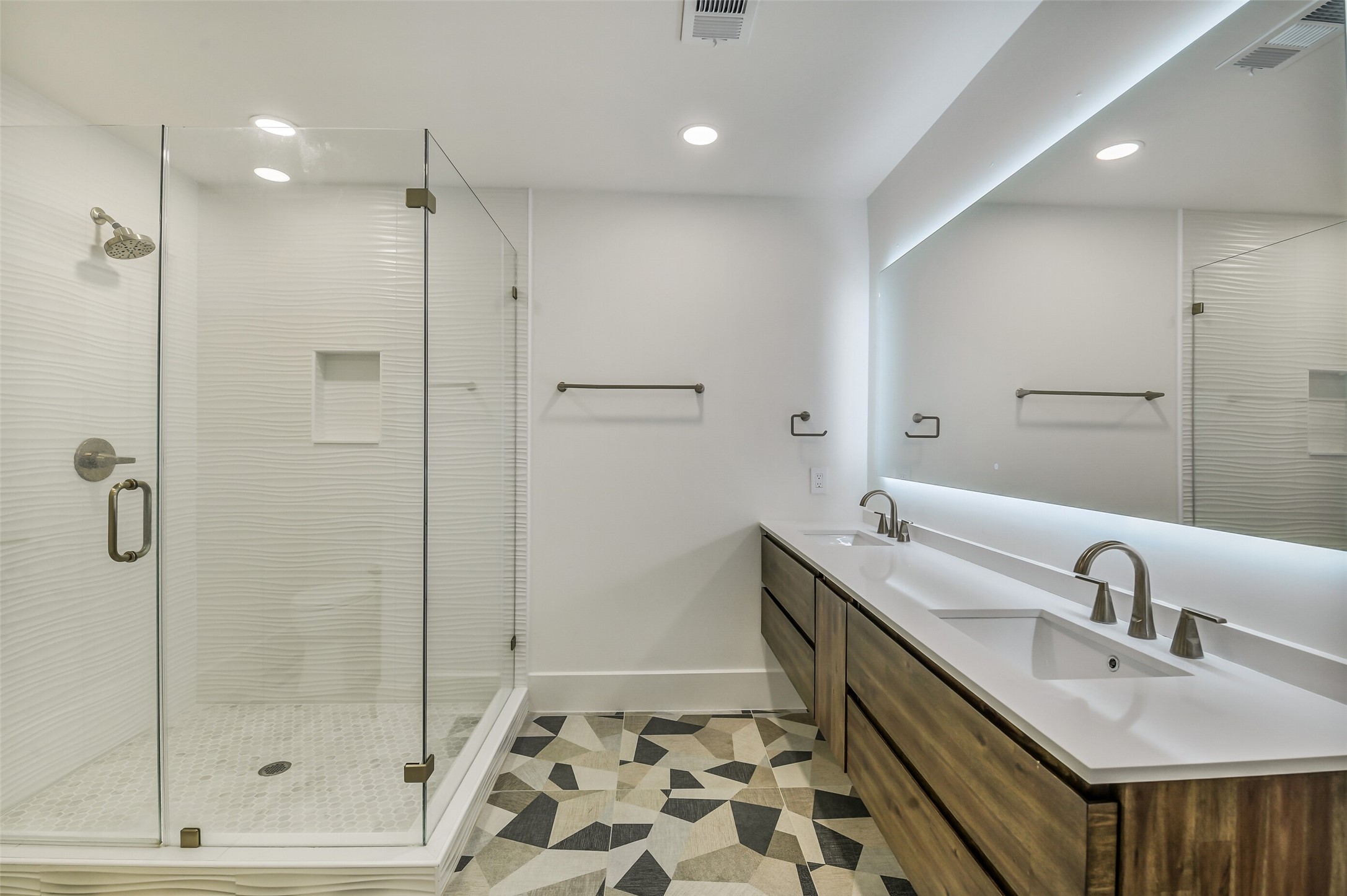 A modern bathroom featuring a glass-enclosed shower with textured tile, a floating dual-sink vanity, and a striking geometric tile floor. A backlit mirror and warm wood cabinetry add a sleek, contemporary finish.
