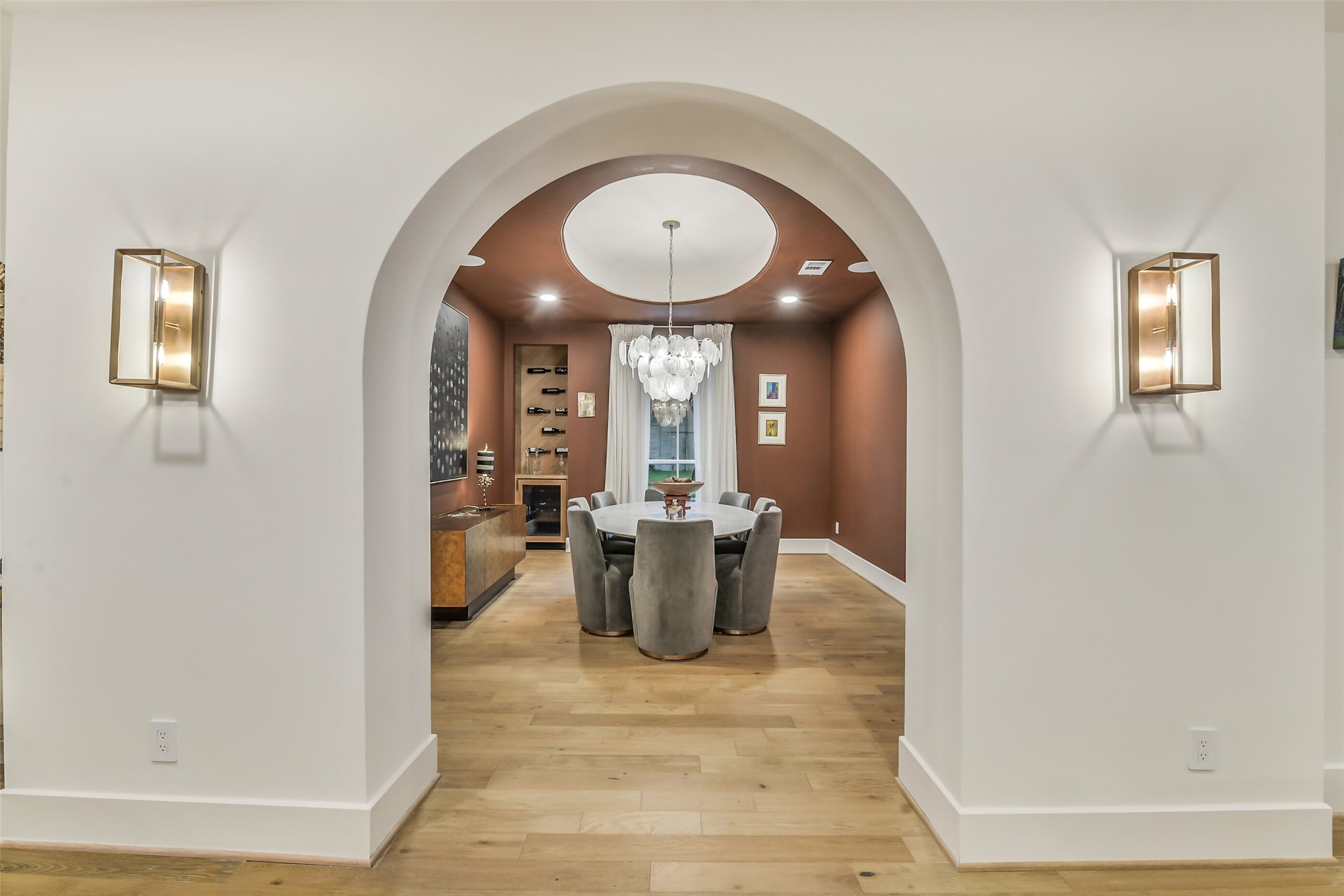 A bold arched entry framed by brass sconces leads into this moody, richly-hued dining room. A statement bubble chandelier hangs from a domed ceiling detail above the round table, while a built-in wine display and custom bar cabinet set the scene for elegant entertaining.