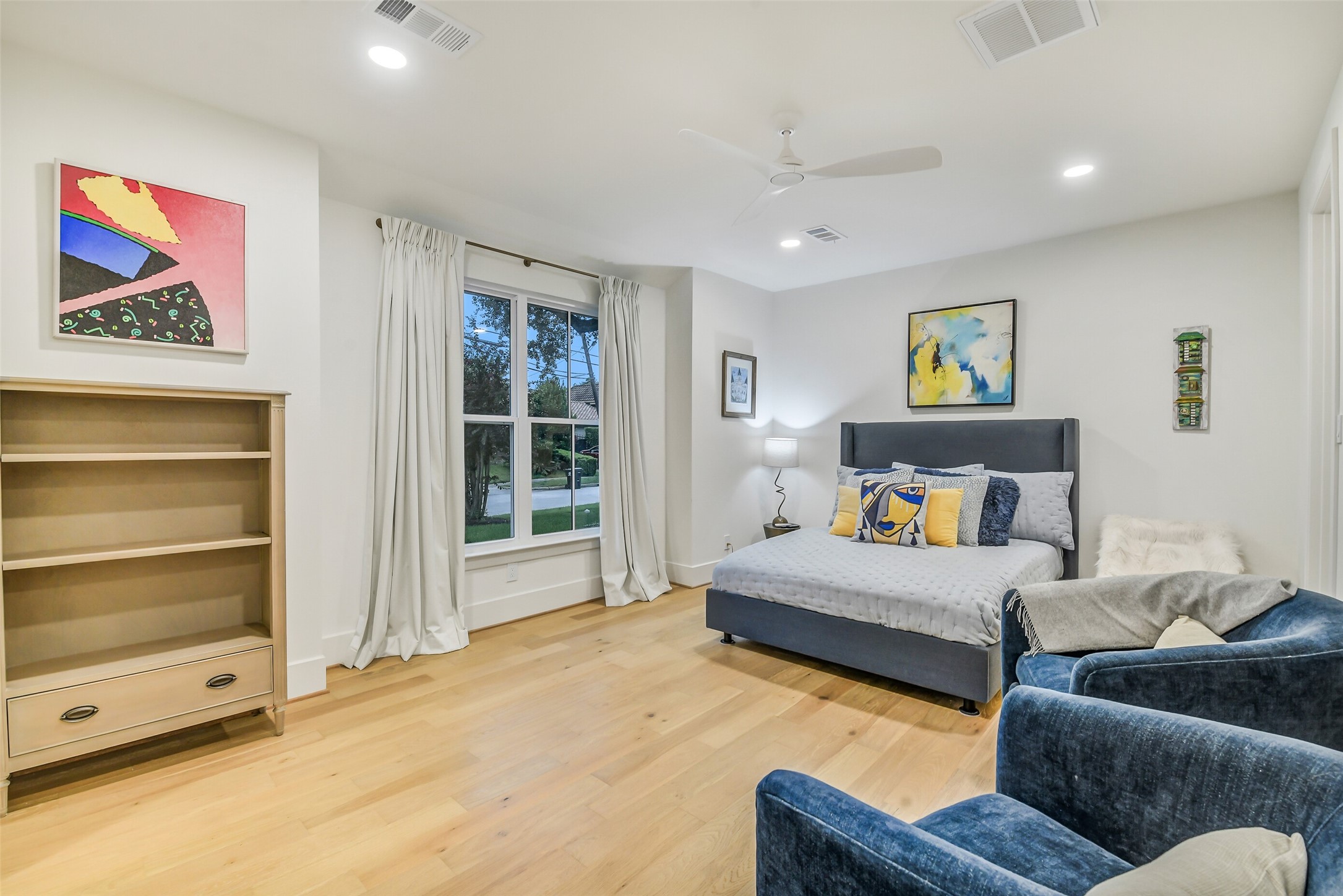 A bright, generously sized secondary bedroom with large windows overlooking the front yard, a ceiling fan, and plenty of room for both a bed and a cozy sitting area. Neutral tones and light wood flooring keep the space airy and adaptable.