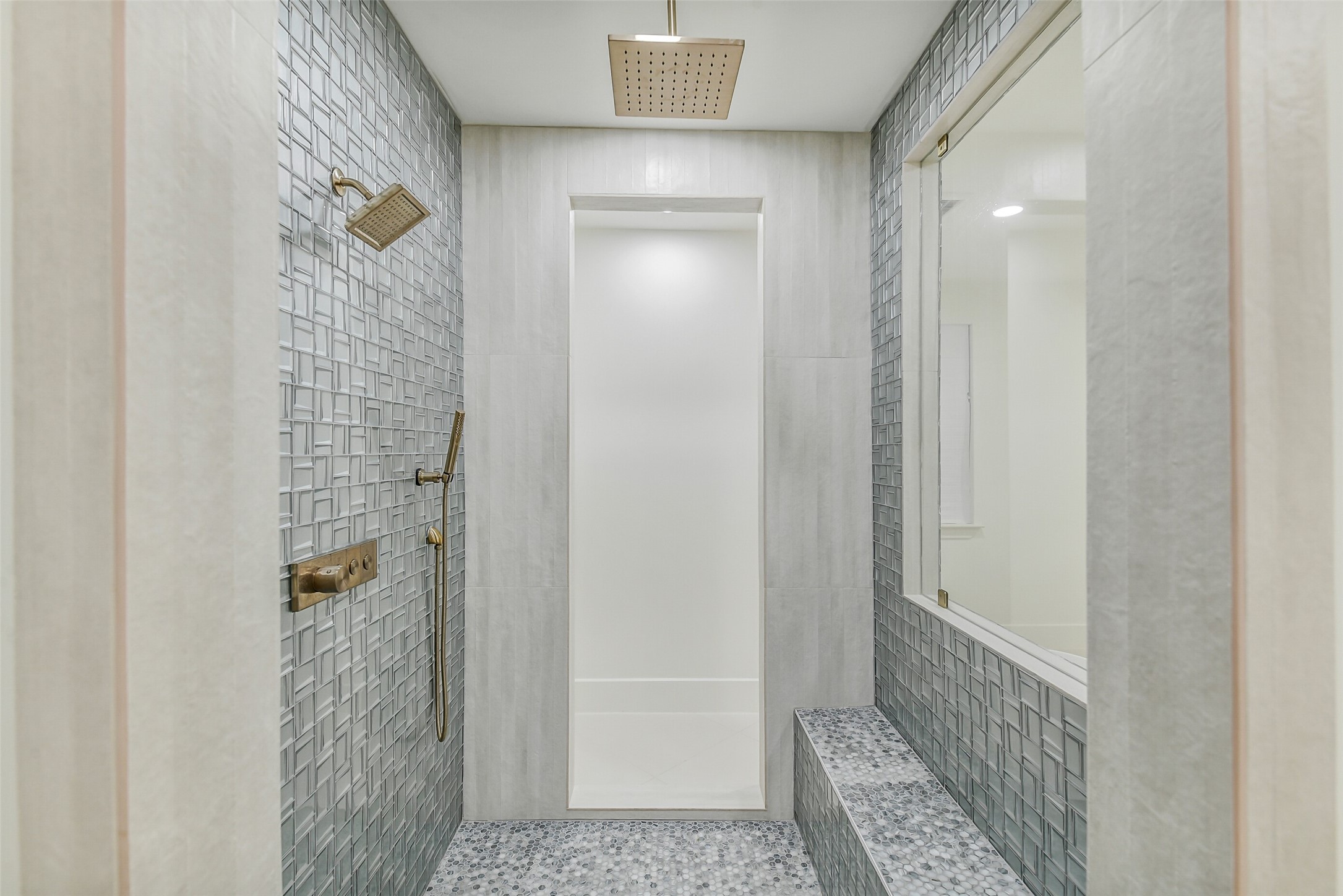 An expansive walk-in shower featuring textured mosaic tile, a built-in bench, and dual showerheads  a rain head overhead plus a handheld sprayer. Brushed brass fixtures and a pebble tile floor add a spa-inspired finishing touch.