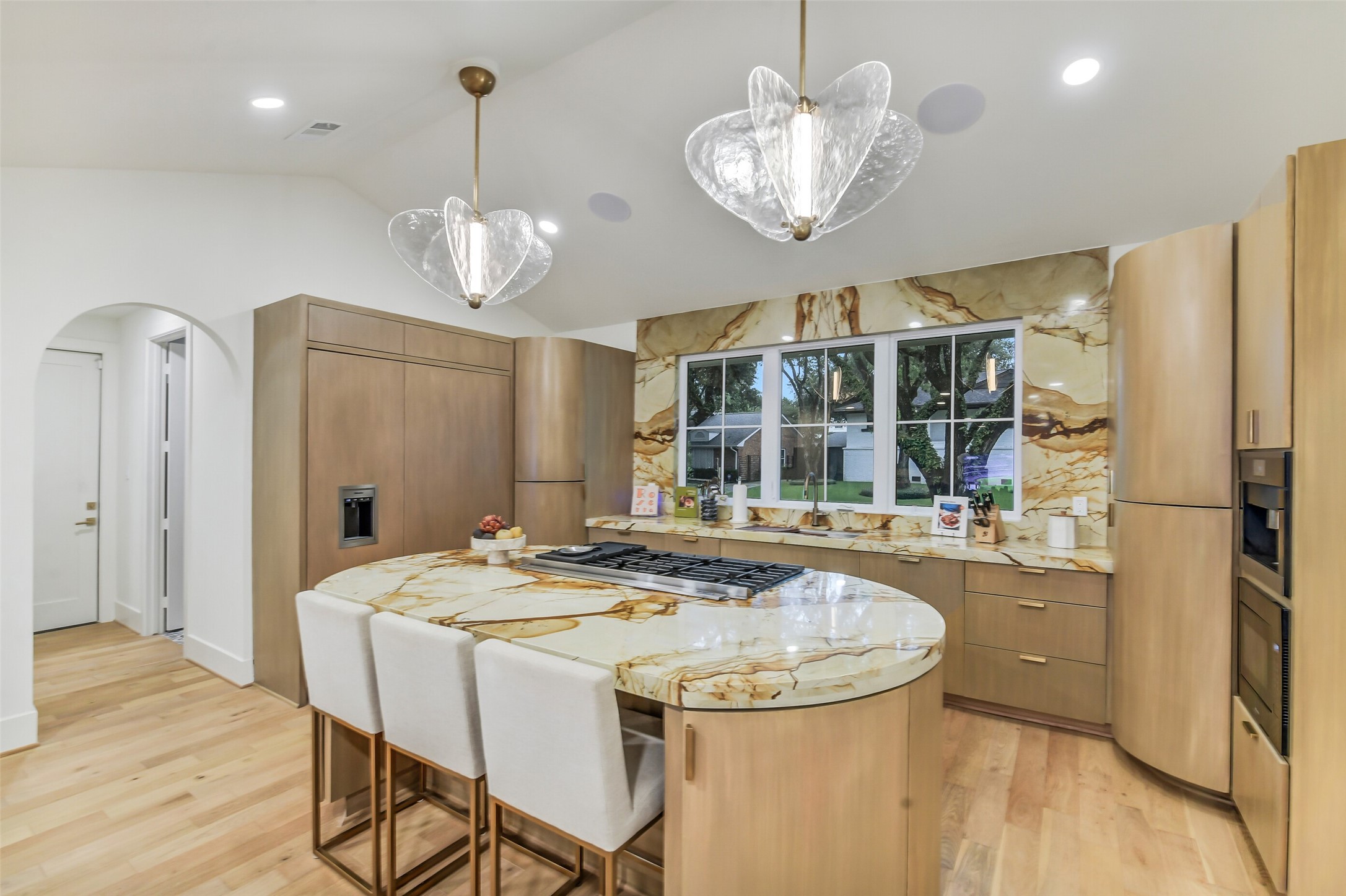 A striking kitchen featuring dramatic stone-slab backsplash, warm wood cabinetry, and an oversized island with bar seating beneath sculptural glass pendant lighting. Paneled-front appliances and a sunny window over the sink complete this elegant, functional space.