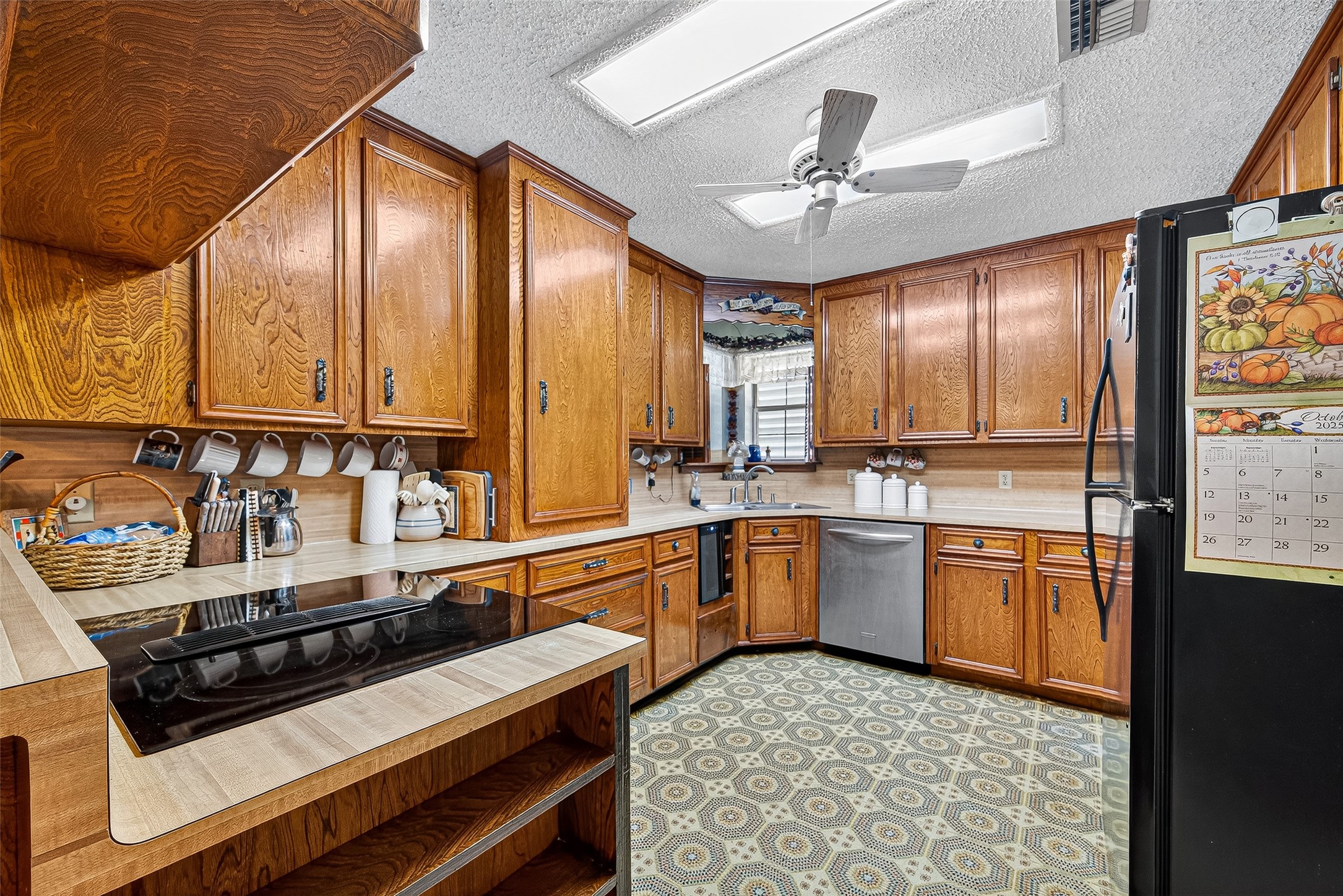 This kitchen features classic wooden cabinetry with ample storage, a modern cooktop, and a stainless steel dishwasher. It has a cozy atmosphere with patterned flooring, ceiling fan, and natural light from a window above the sink.